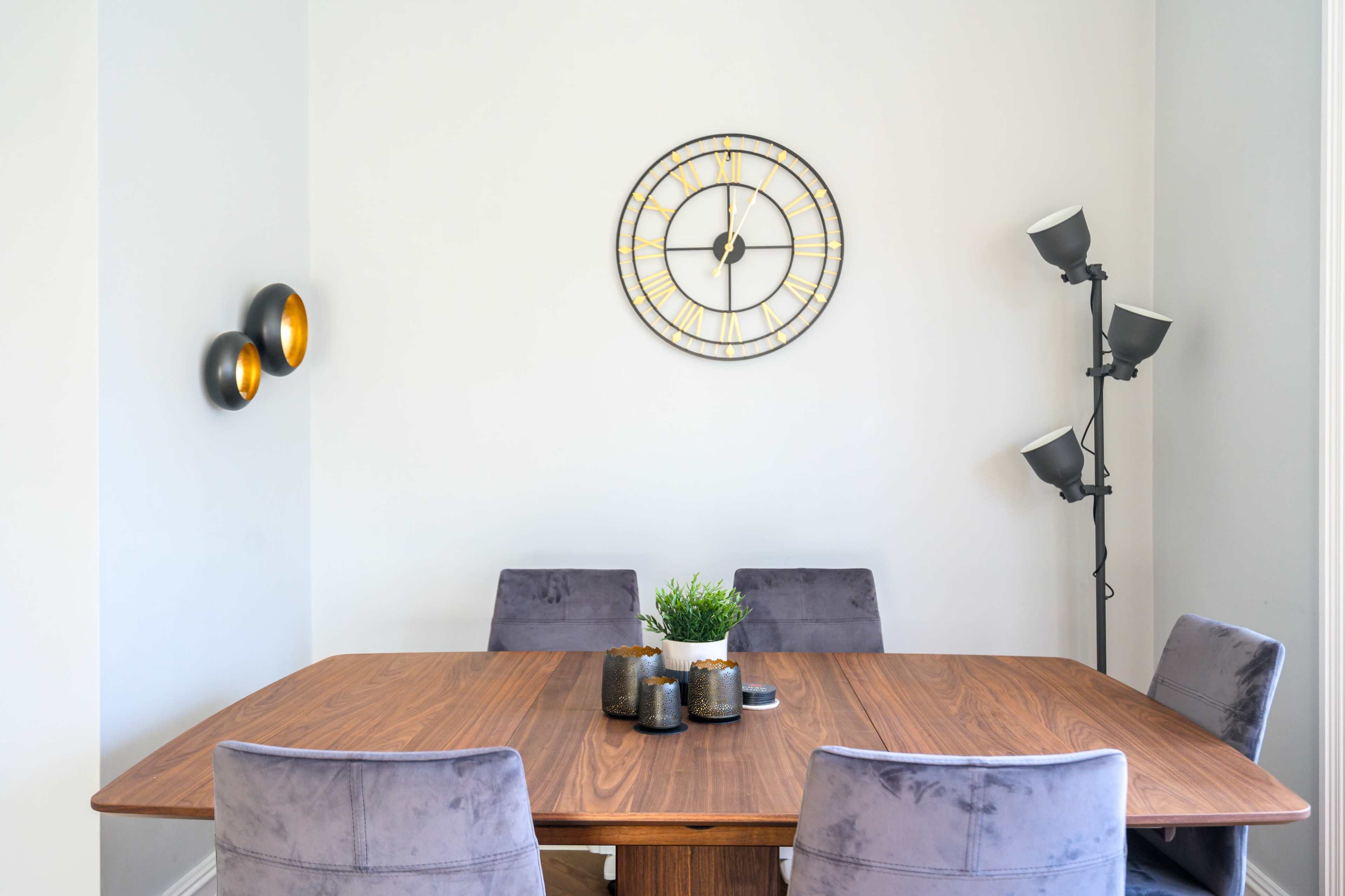 A wooden dining table with four upholstered chairs is positioned in a simple room featuring a wall clock and a floor lamp.