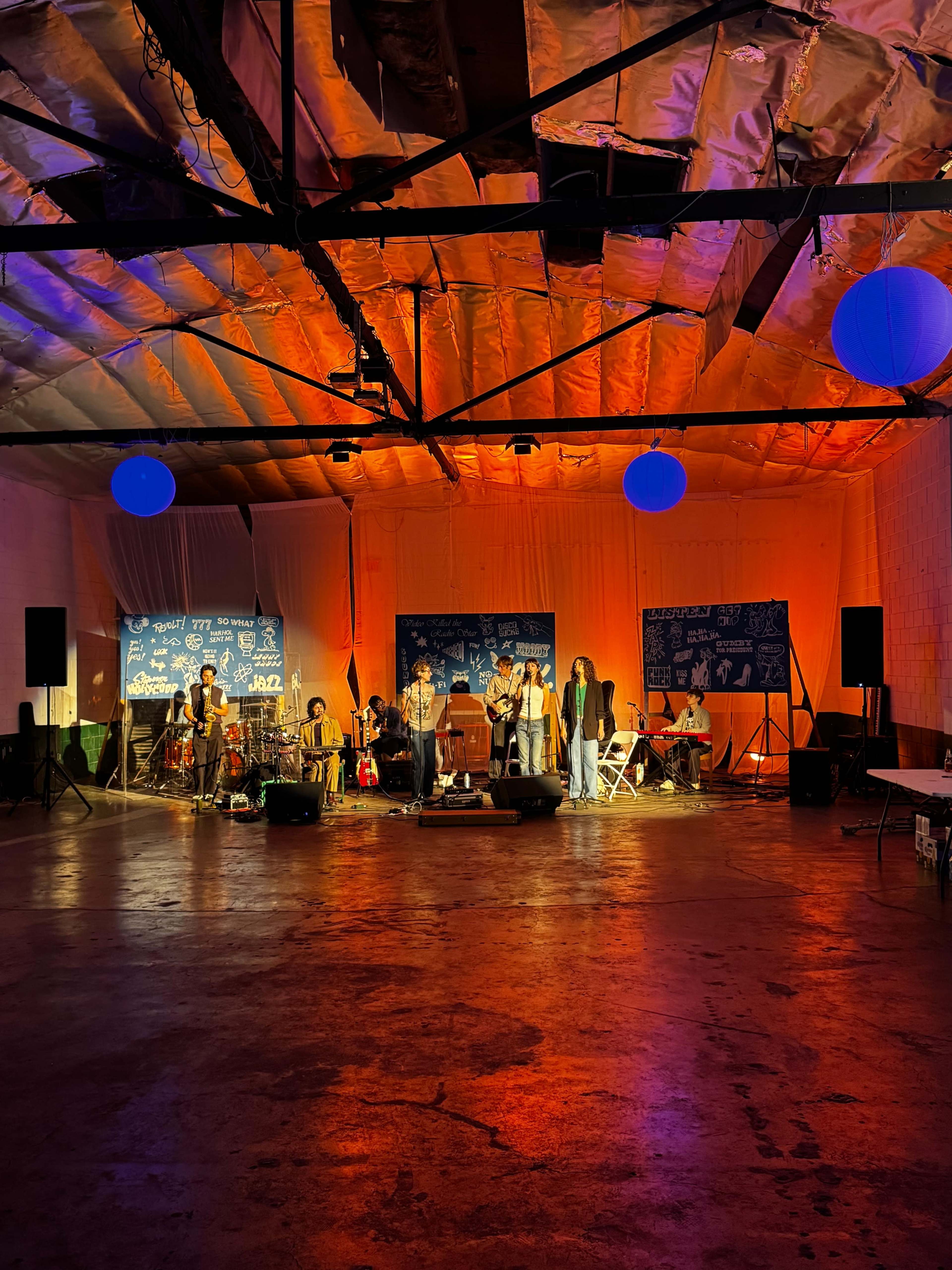A band performs on stage in a dimly lit warehouse, with colorful lights illuminating the space and an audience watching.