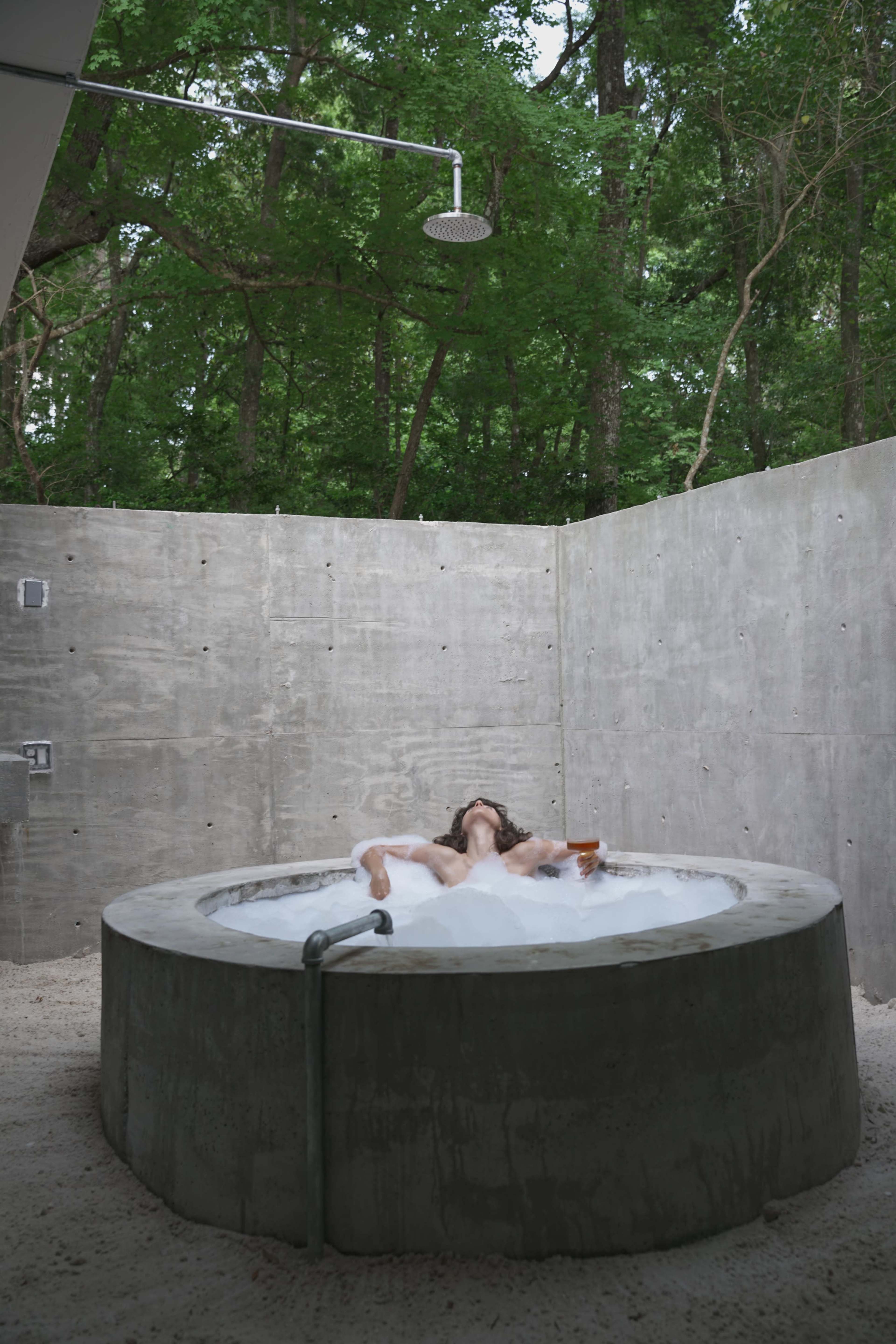 A person relaxes in a circular concrete hot tub surrounded by concrete walls and lush greenery.