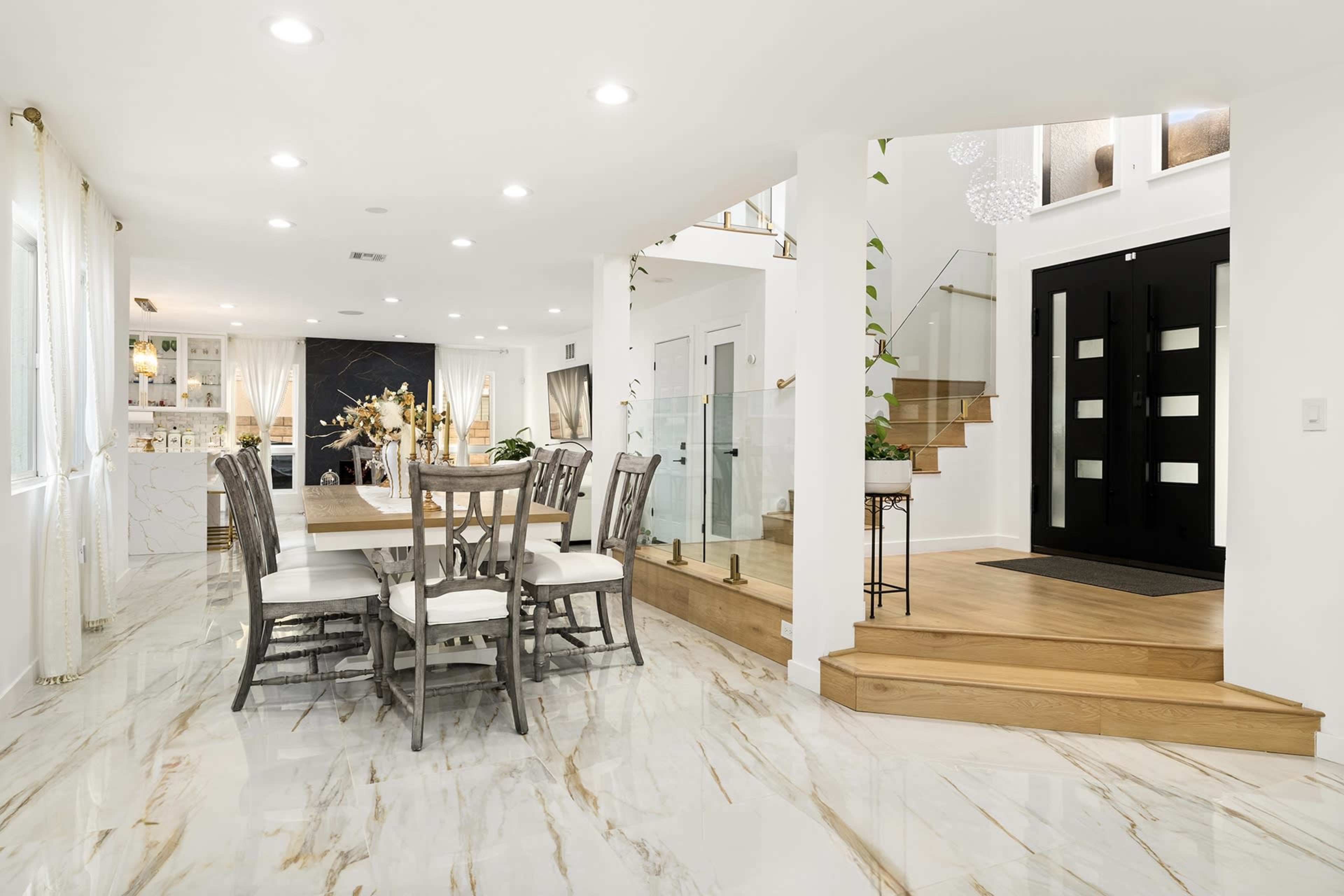 The image shows a spacious modern dining area with a wooden table and chairs, adjacent to a staircase leading to another level and a black front door.