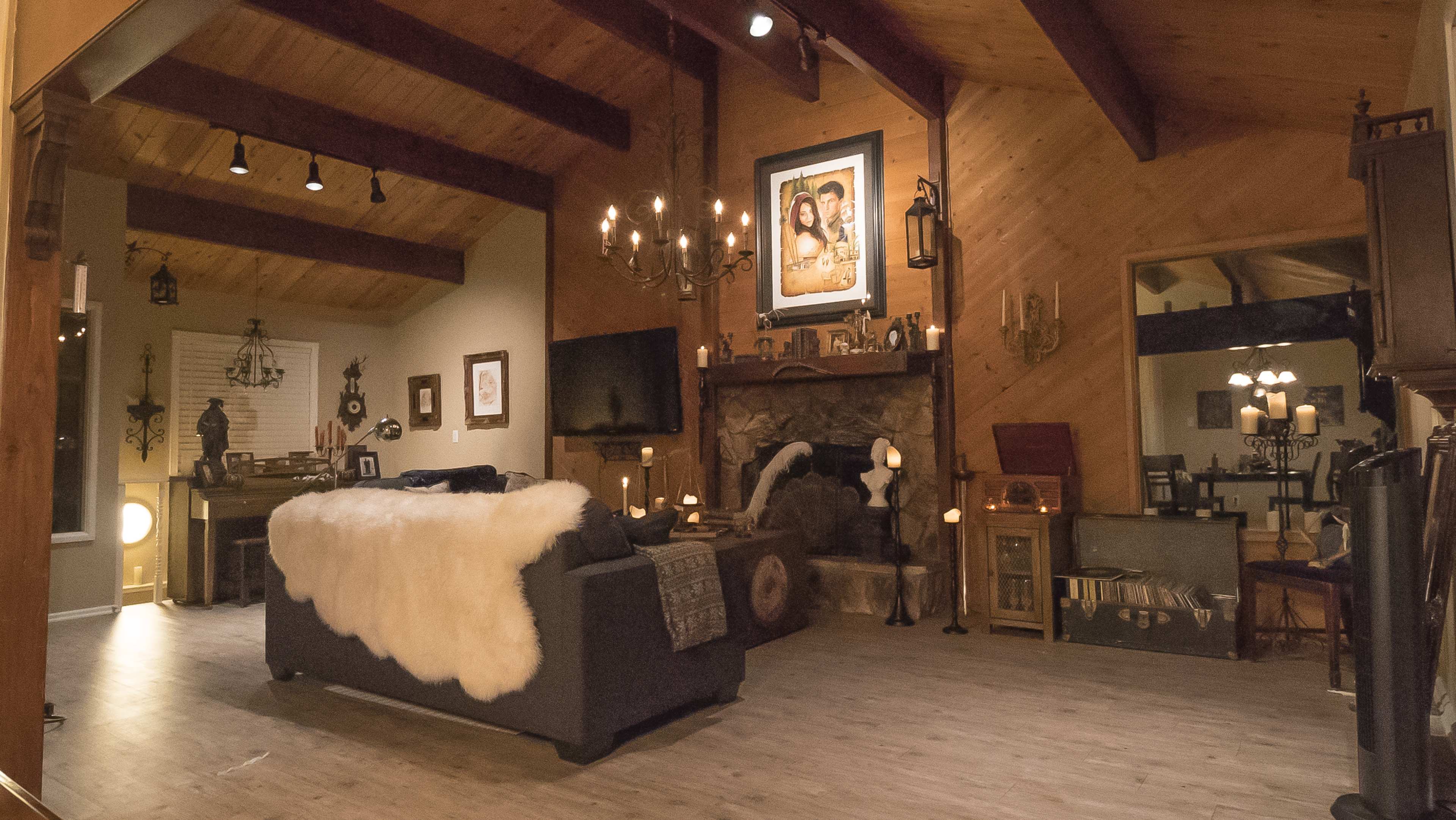 The scene features a cozy living room with rustic wooden beams, a stone fireplace adorned with candles, and a large fur throw draped over a dark sofa.