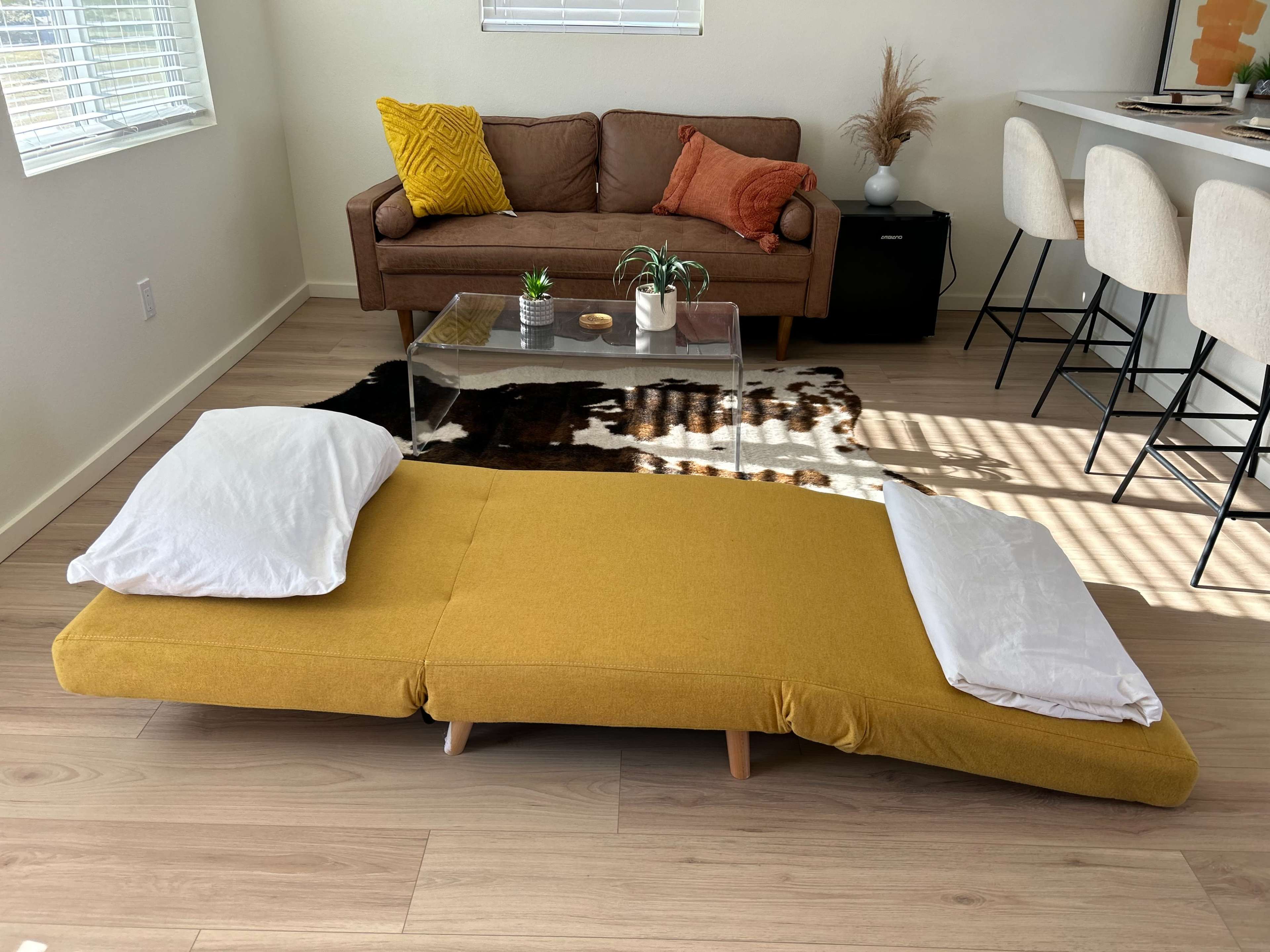 A yellow, low-profile futon mattress with two pillows is placed on a light wooden floor in a modern living room featuring a brown couch and a glass coffee table.
