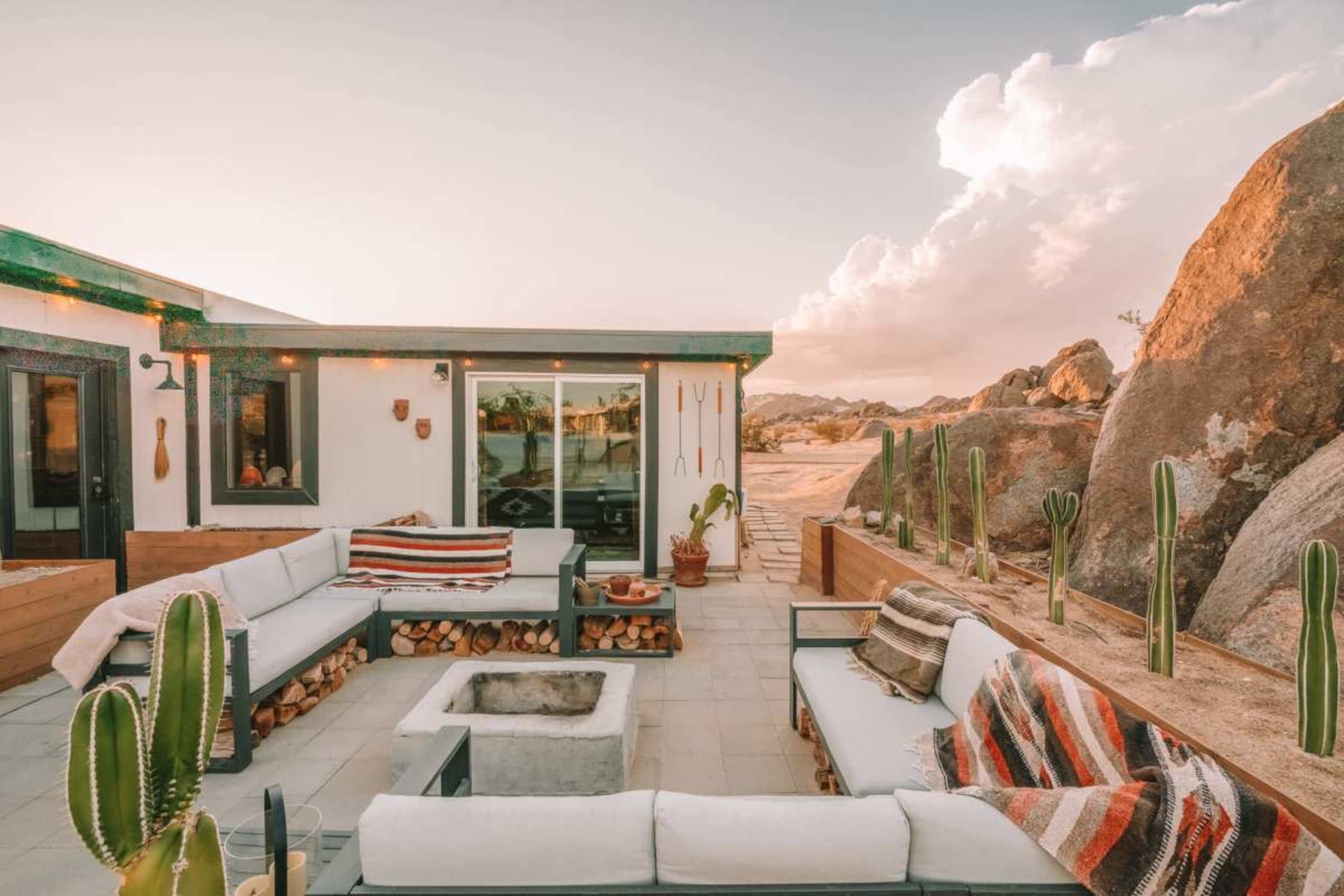 The image shows a modern outdoor seating area surrounded by boulders and cacti, featuring a fire pit and a view of the desert landscape.