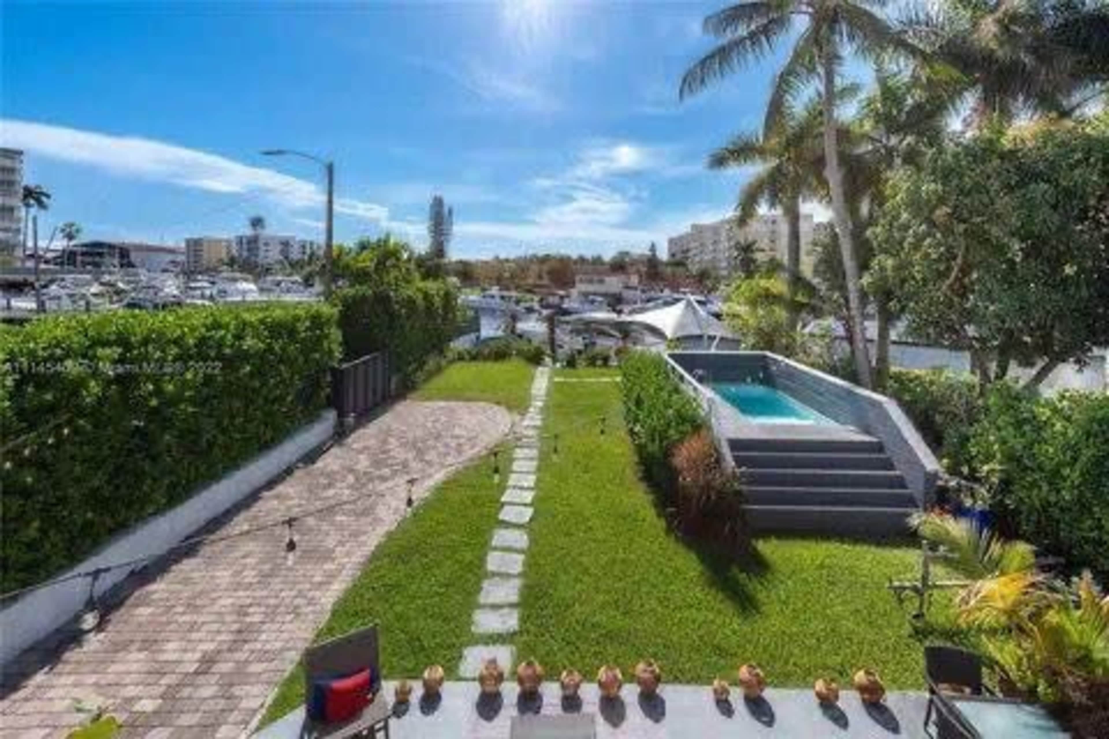 The image shows a landscaped backyard leading to a small swimming pool and a view of boats in a marina under a clear sky.