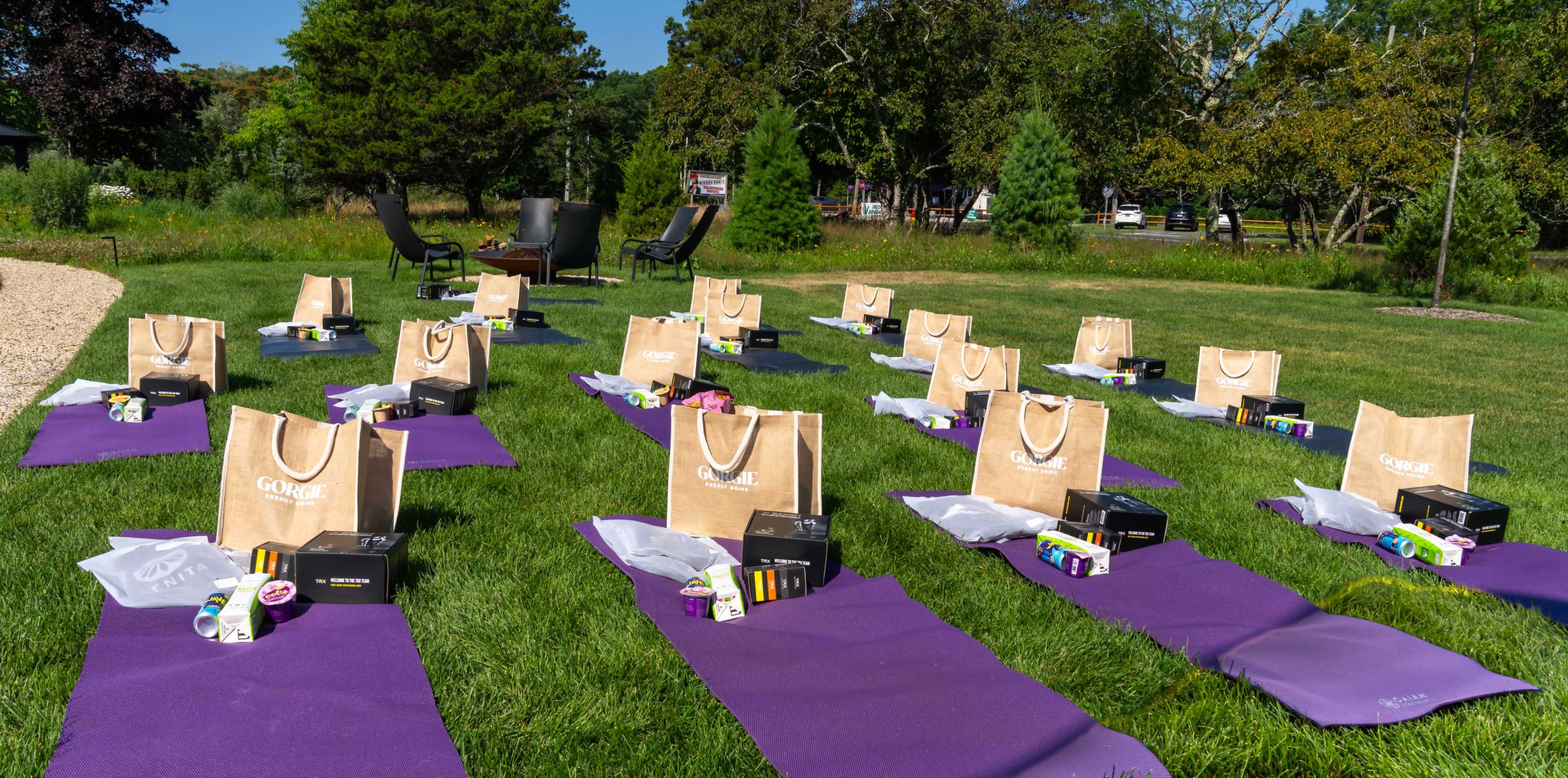 The image shows several yoga mats arranged in rows on a grassy area, each with a tote bag and various items placed on them.