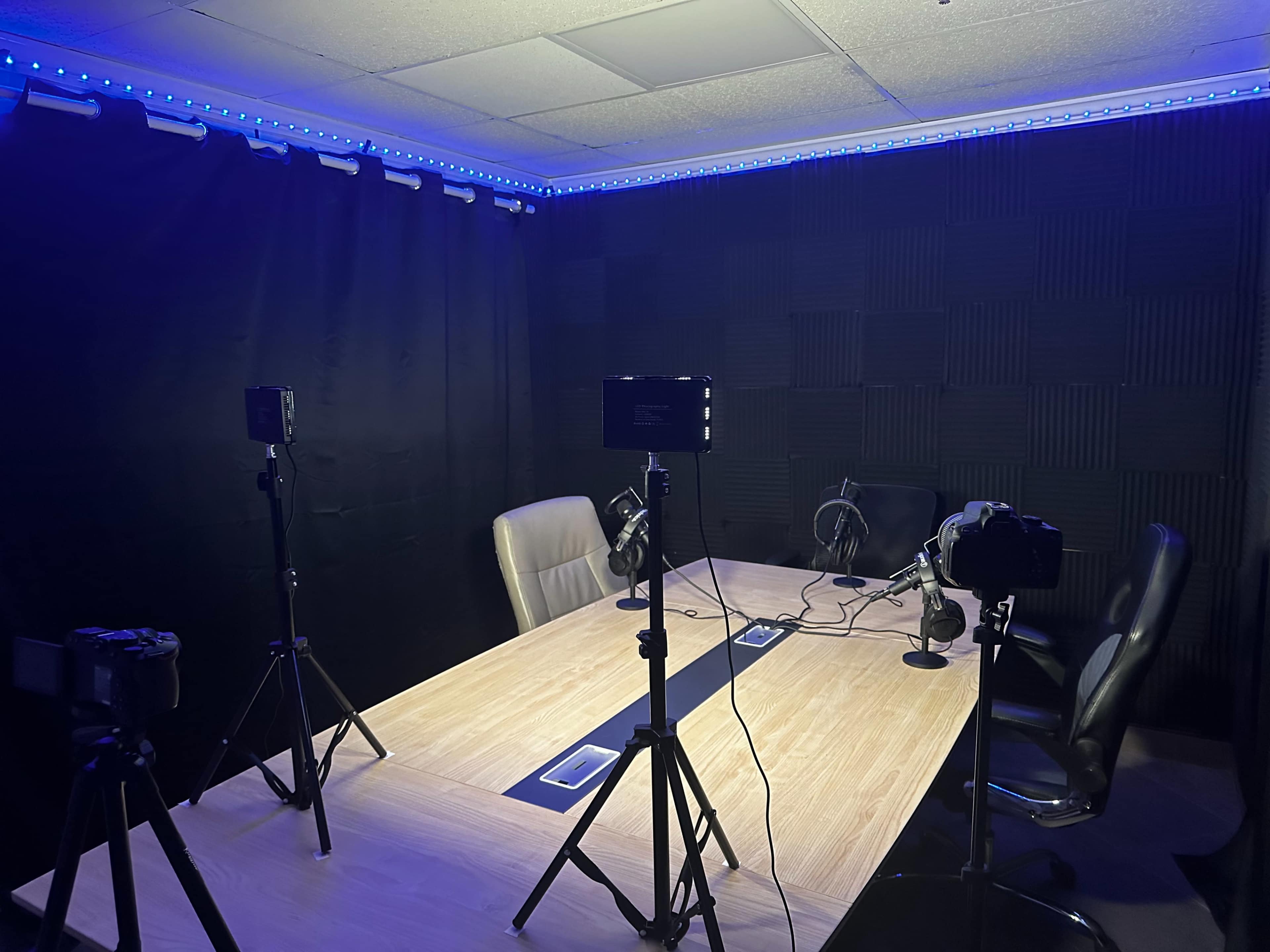 A recording studio features a large table surrounded by chairs, with multiple cameras and microphones set up on tripods, and blue lighting along the ceiling.