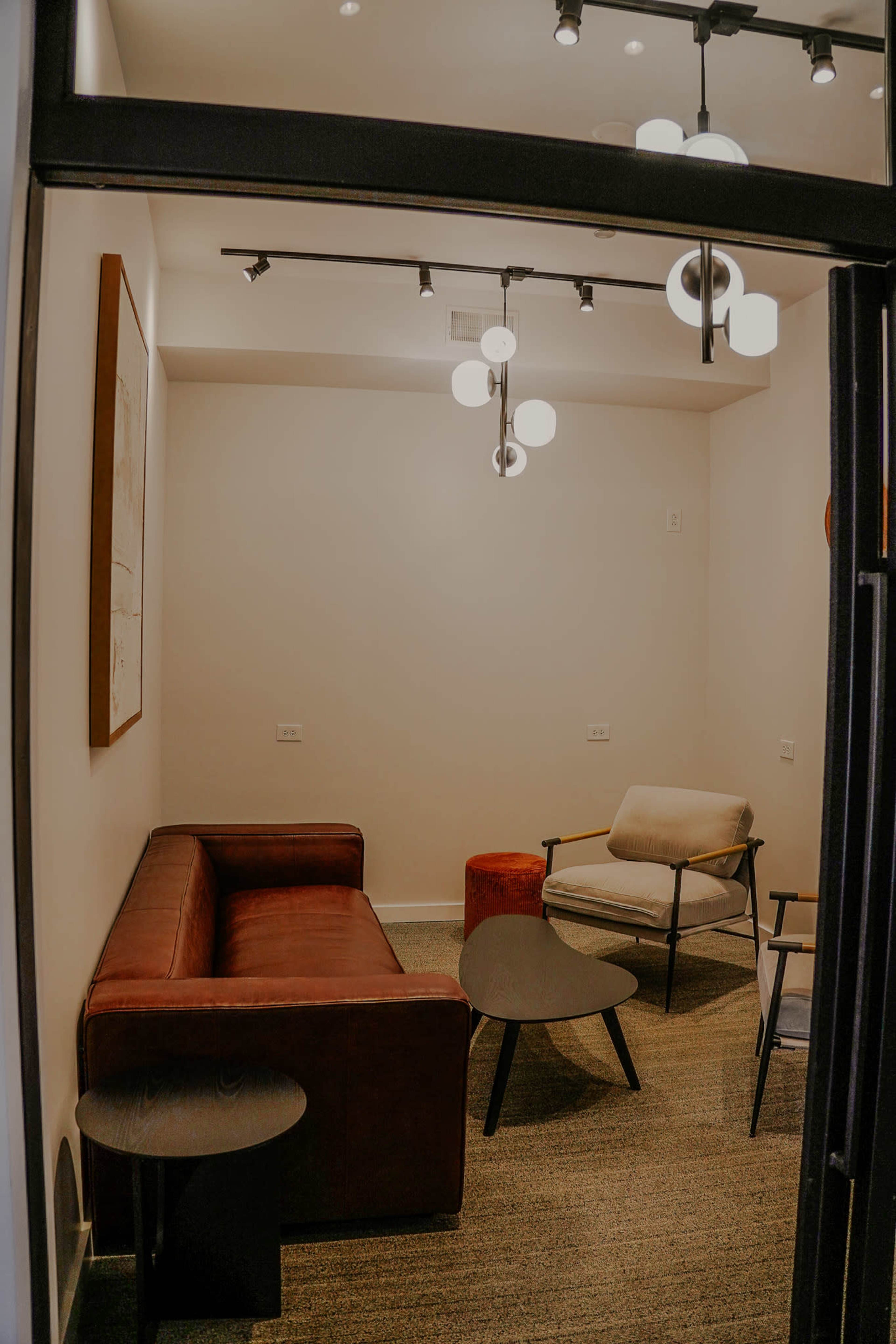 The image shows a small, modern lounge area with a brown leather sofa, a beige armchair, a circular coffee table, and minimalistic lighting fixtures.