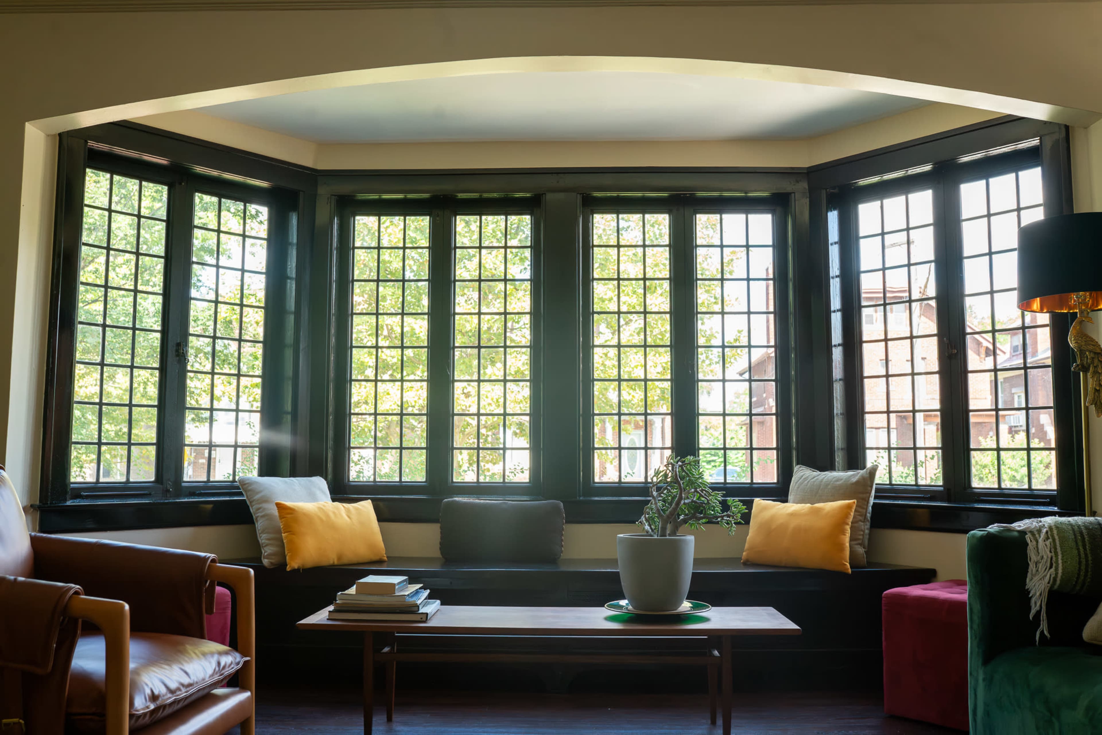The image shows a sunny bay window area with multiple grid-patterned windows, a black window seat adorned with yellow cushions, and a small table displaying a plant.