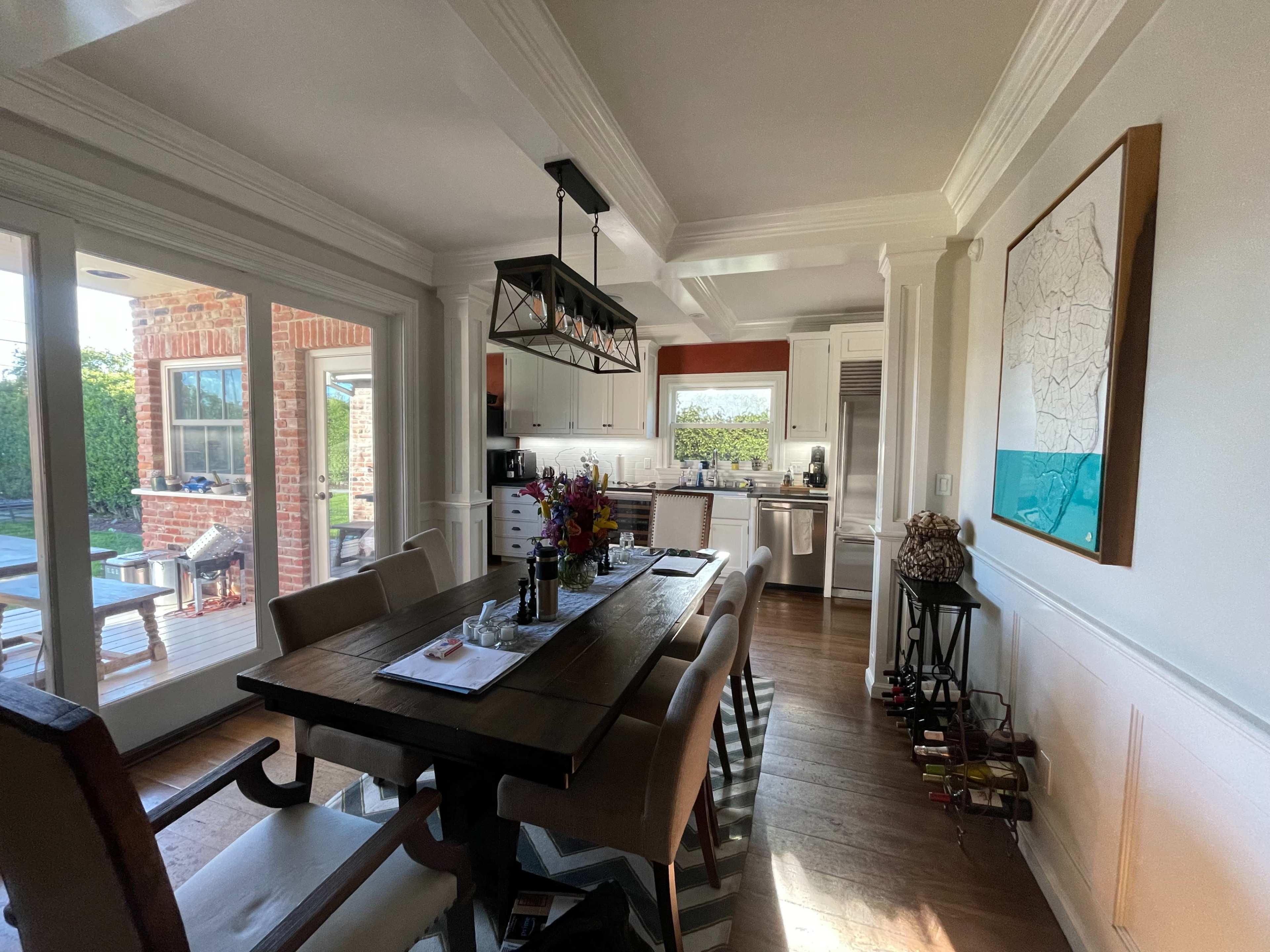 A dining area featuring a wooden table with chairs, surrounded by large windows that provide light to a kitchen in the background.