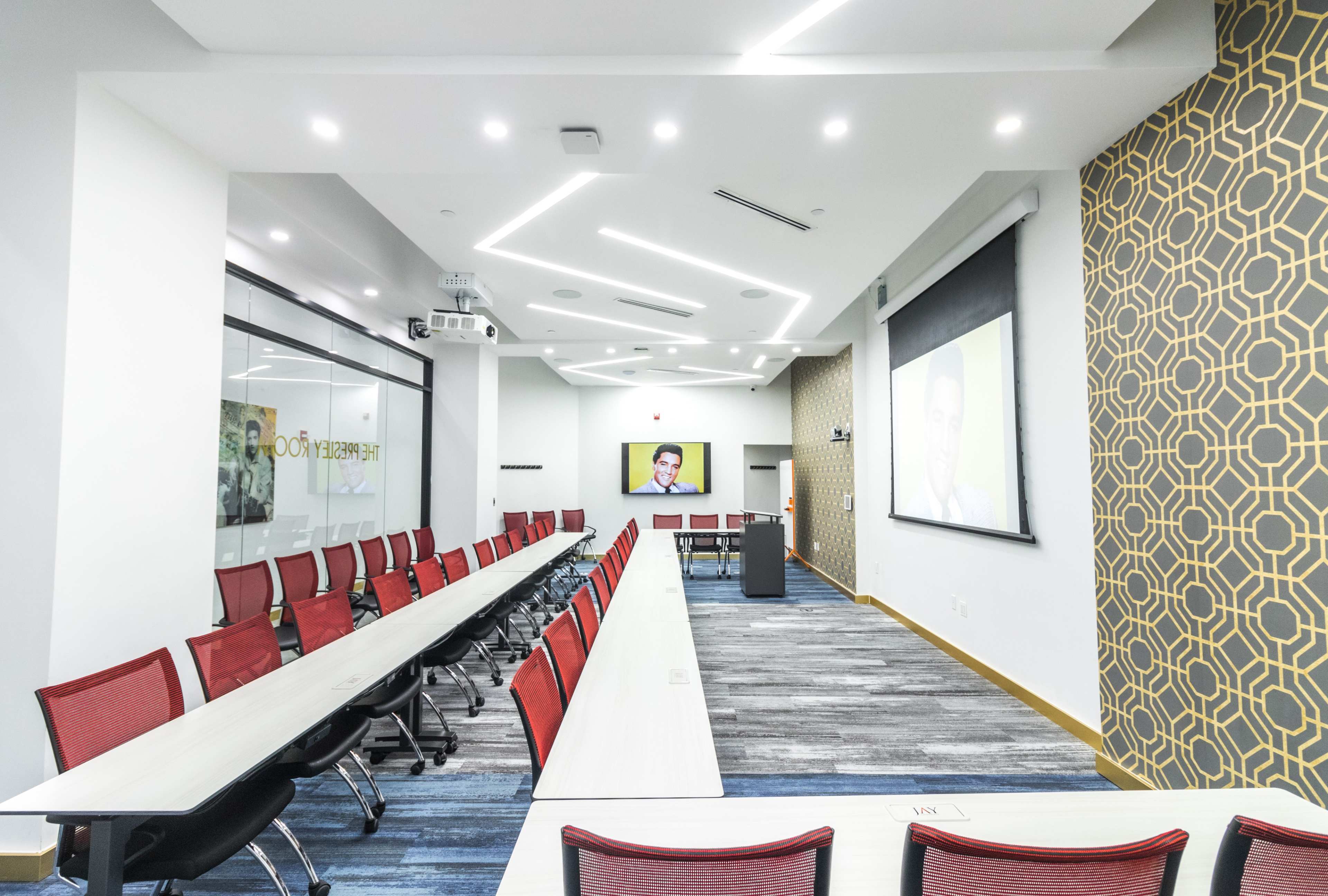 The image shows a modern conference room with a long table, red chairs, a projection screen, and a wall featuring a geometric design.
