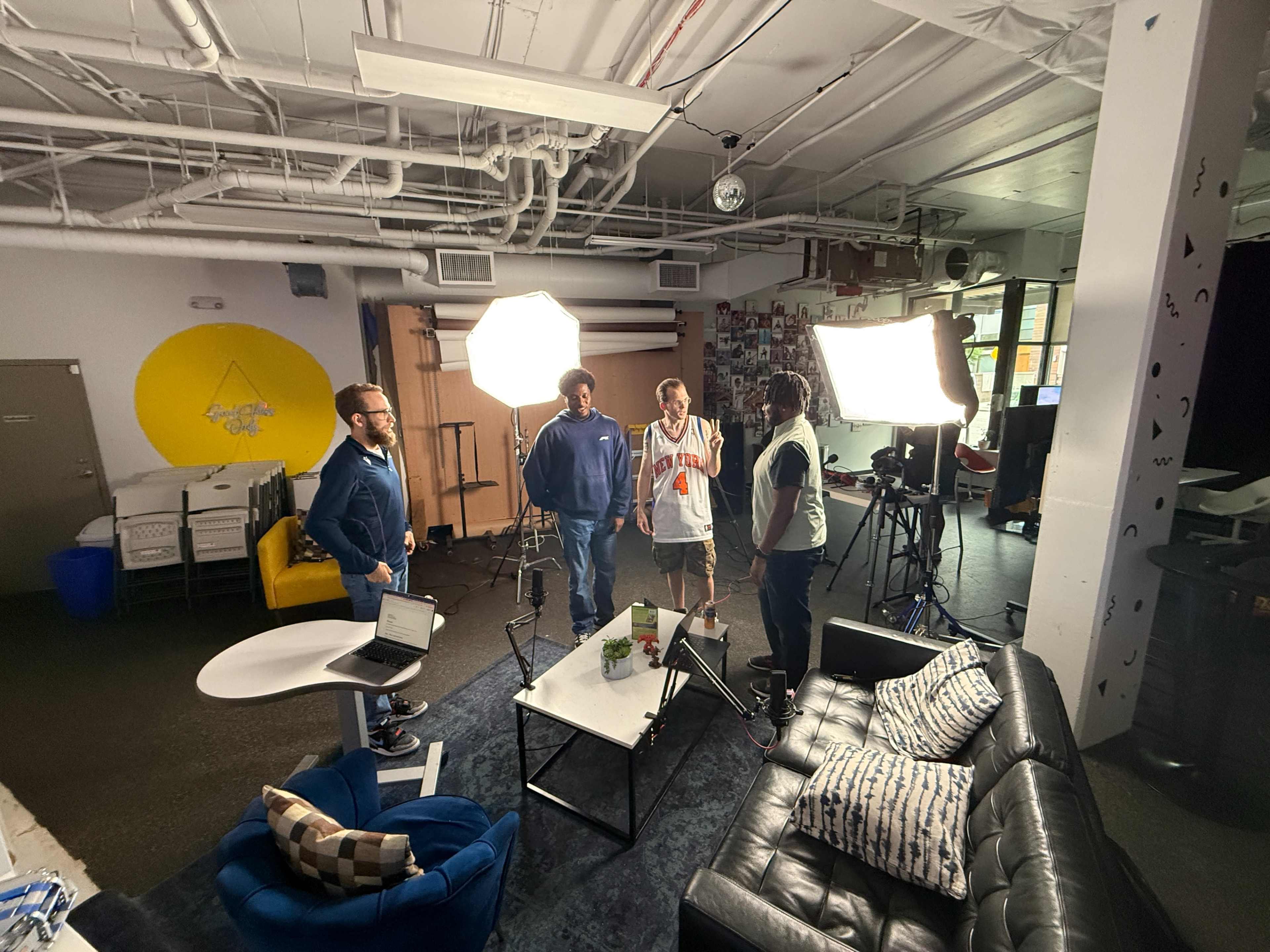 A group of four people is engaged in conversation in a studio setting, with bright lights illuminating the area and a couch in the foreground.