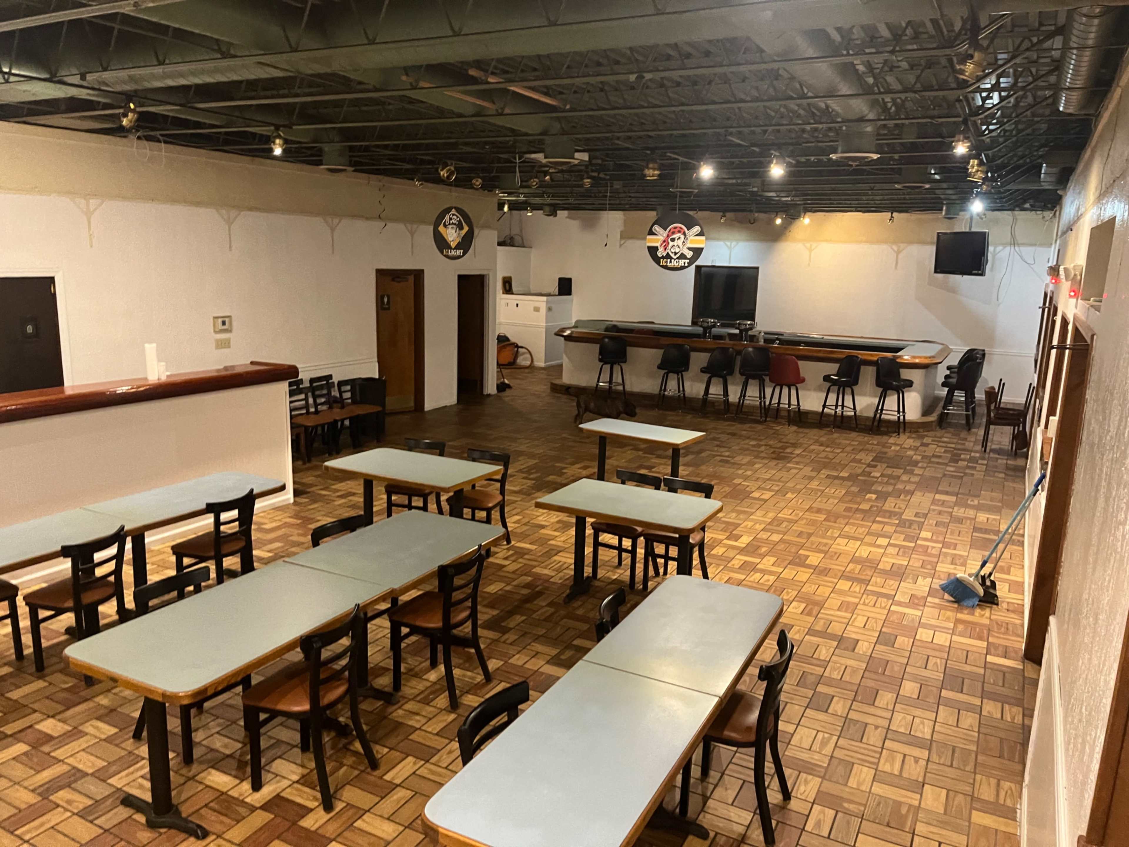 The image shows an empty room with multiple tables and chairs arranged in a grid pattern, a bar area at the back, and various lighting fixtures on the ceiling.