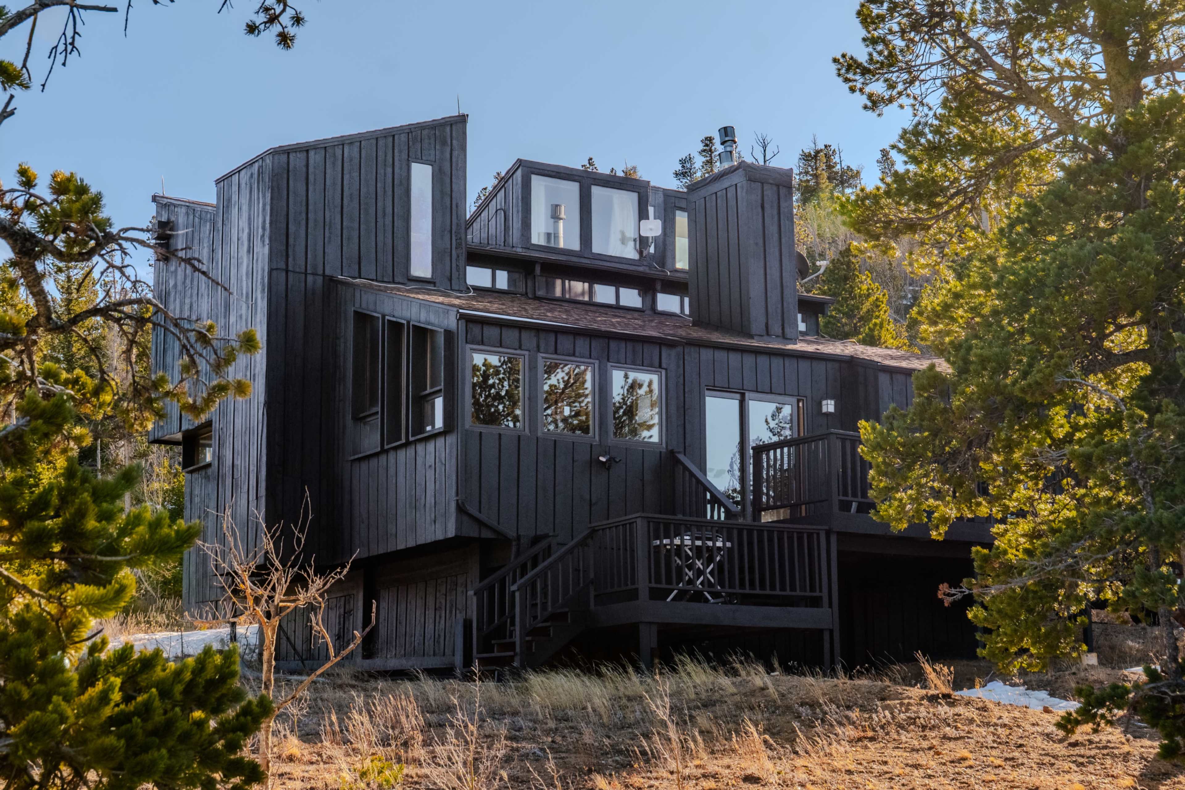 The image shows a modern, dark wood house set among trees on a hillside.