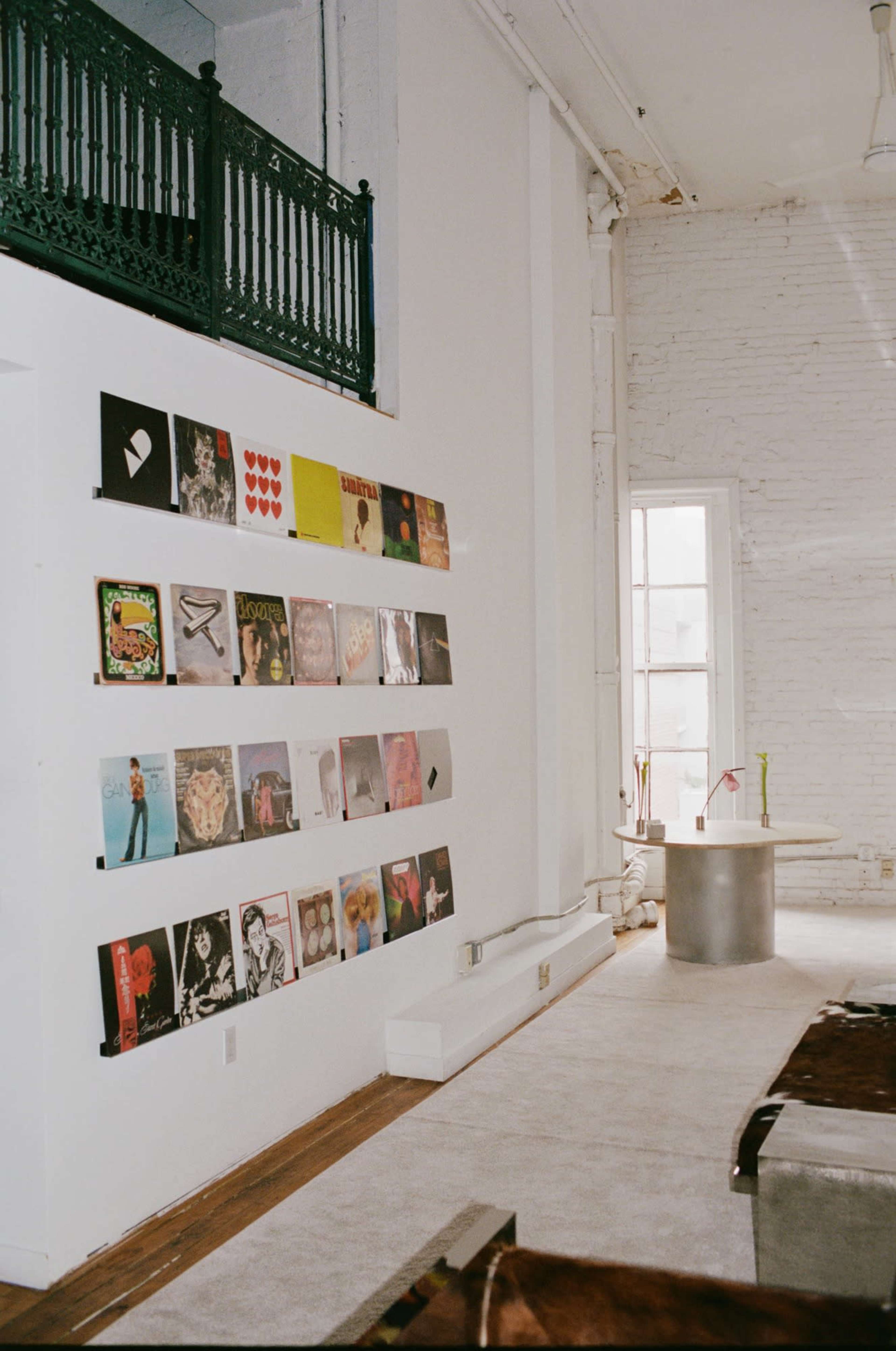 The image depicts a minimalist interior space featuring a wall display of various vinyl record covers, with a round table in the foreground.