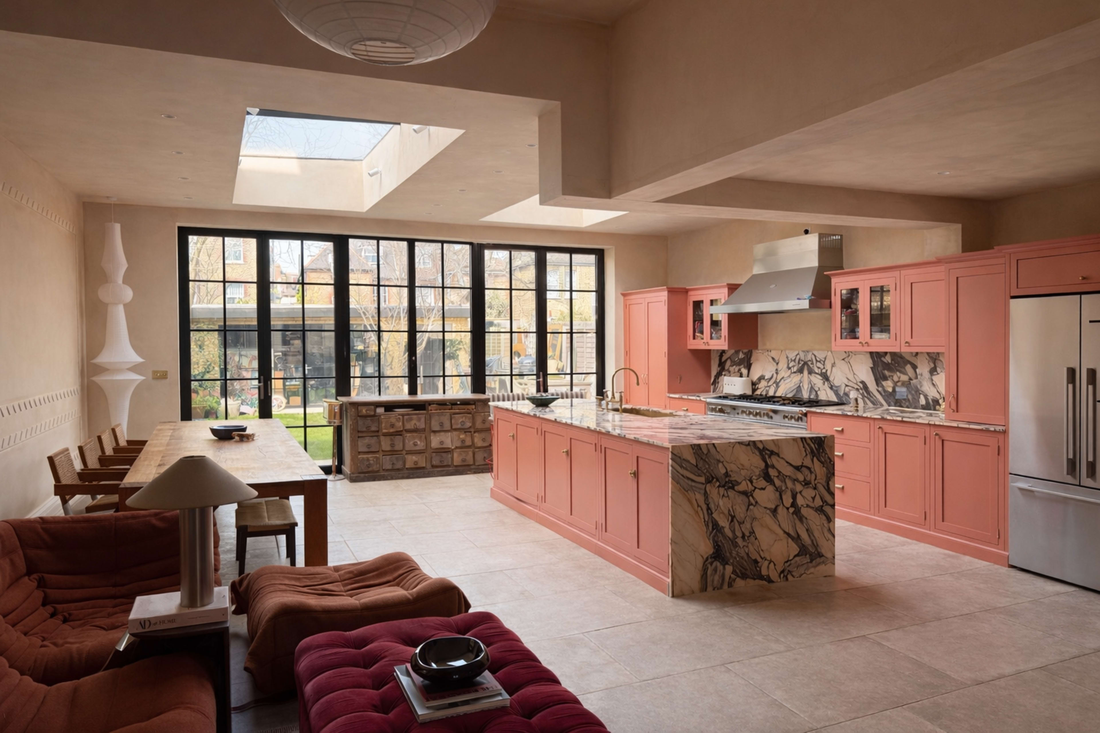 The image shows a modern kitchen with pink cabinetry, a marble countertop, and large windows that open to a garden area.