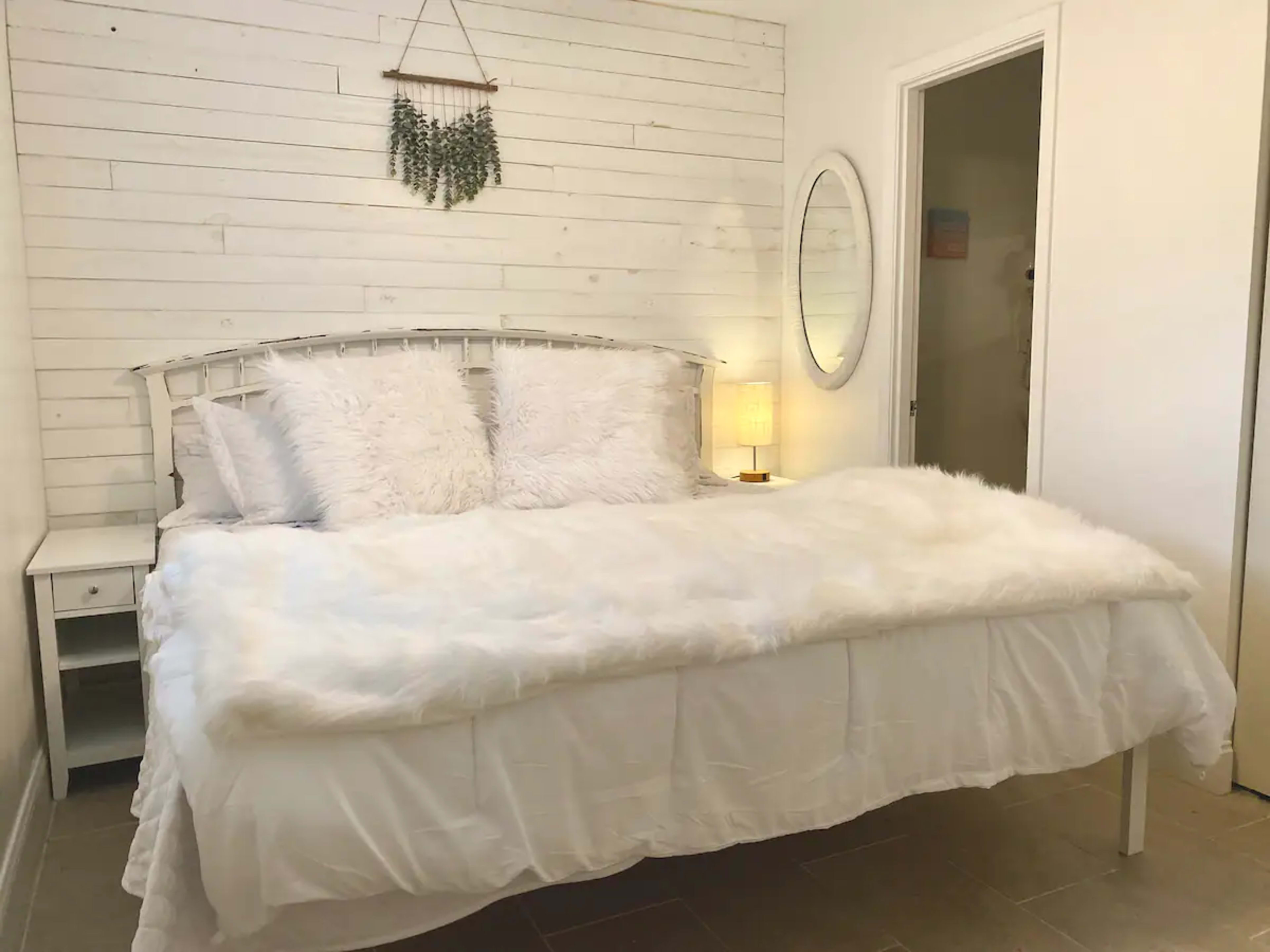 A white bed with fluffy pillows and a fur throw is positioned in a room with whitewashed walls and a round mirror.