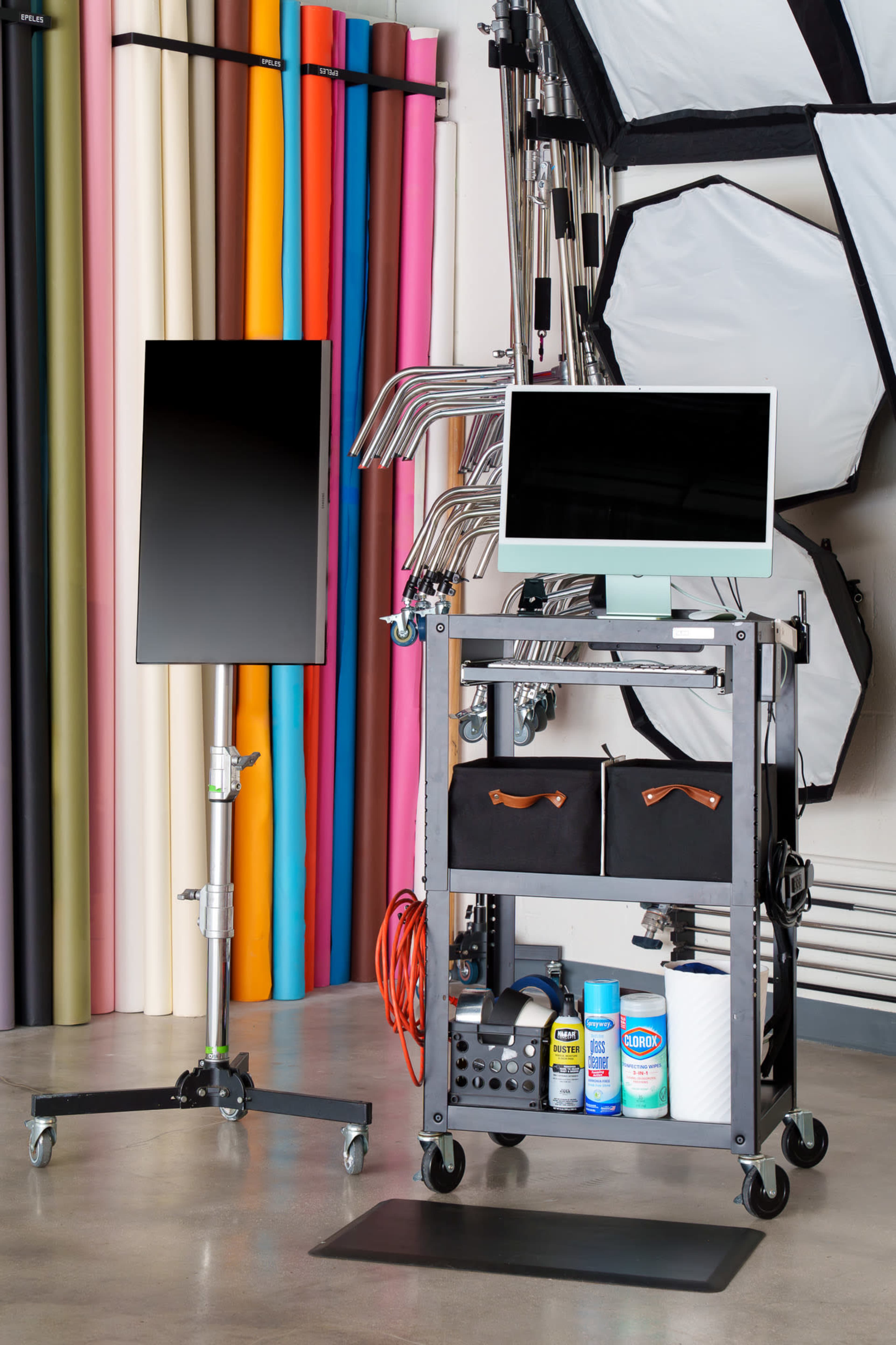The image shows a photography studio setup featuring a rolling cart with a computer monitor and storage bins, alongside a light stand and a backdrop of colorful paper rolls in the background.