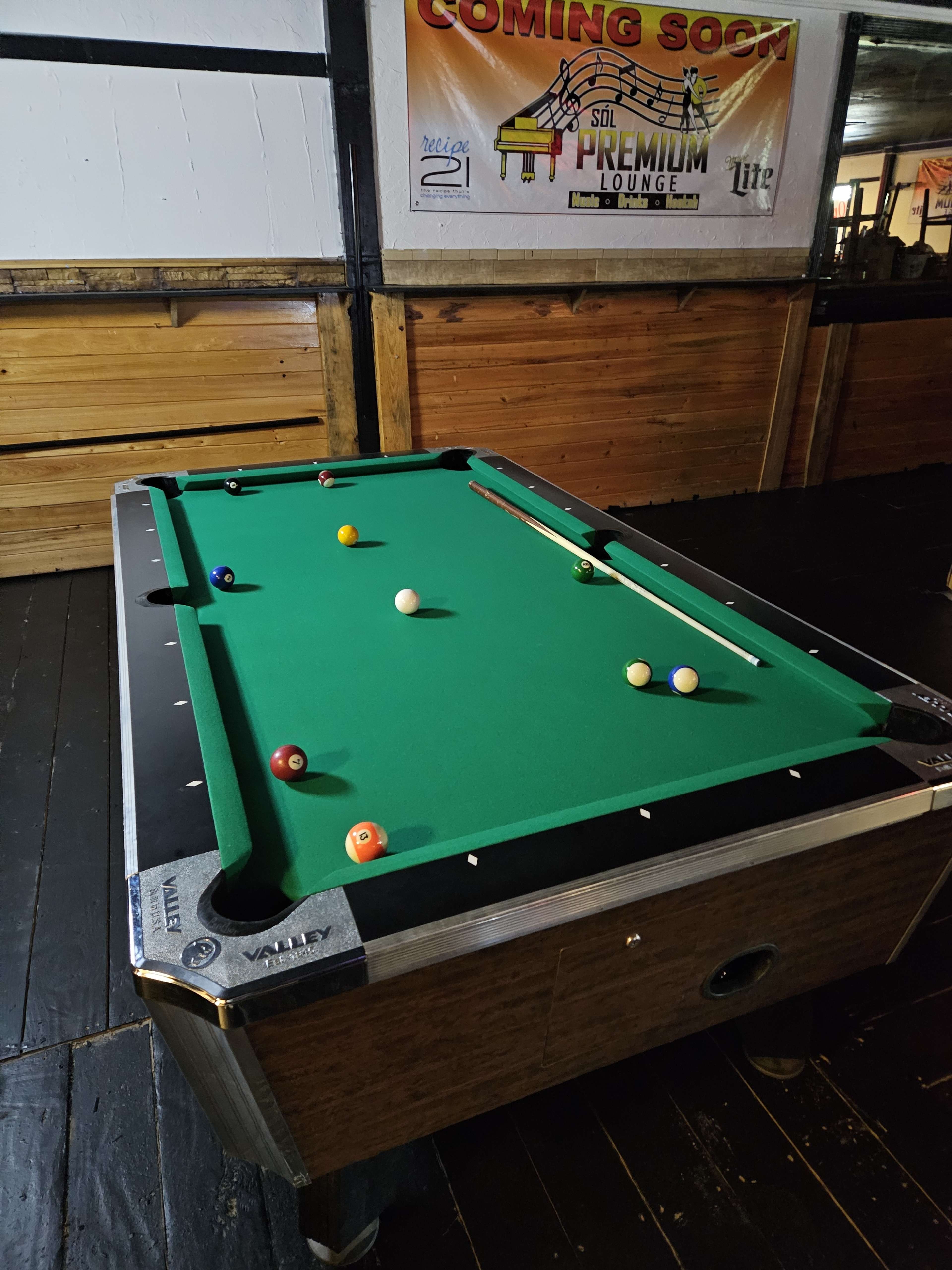 A pool table with a green felt surface and several billiard balls scattered across it is positioned in a dimly lit lounge.