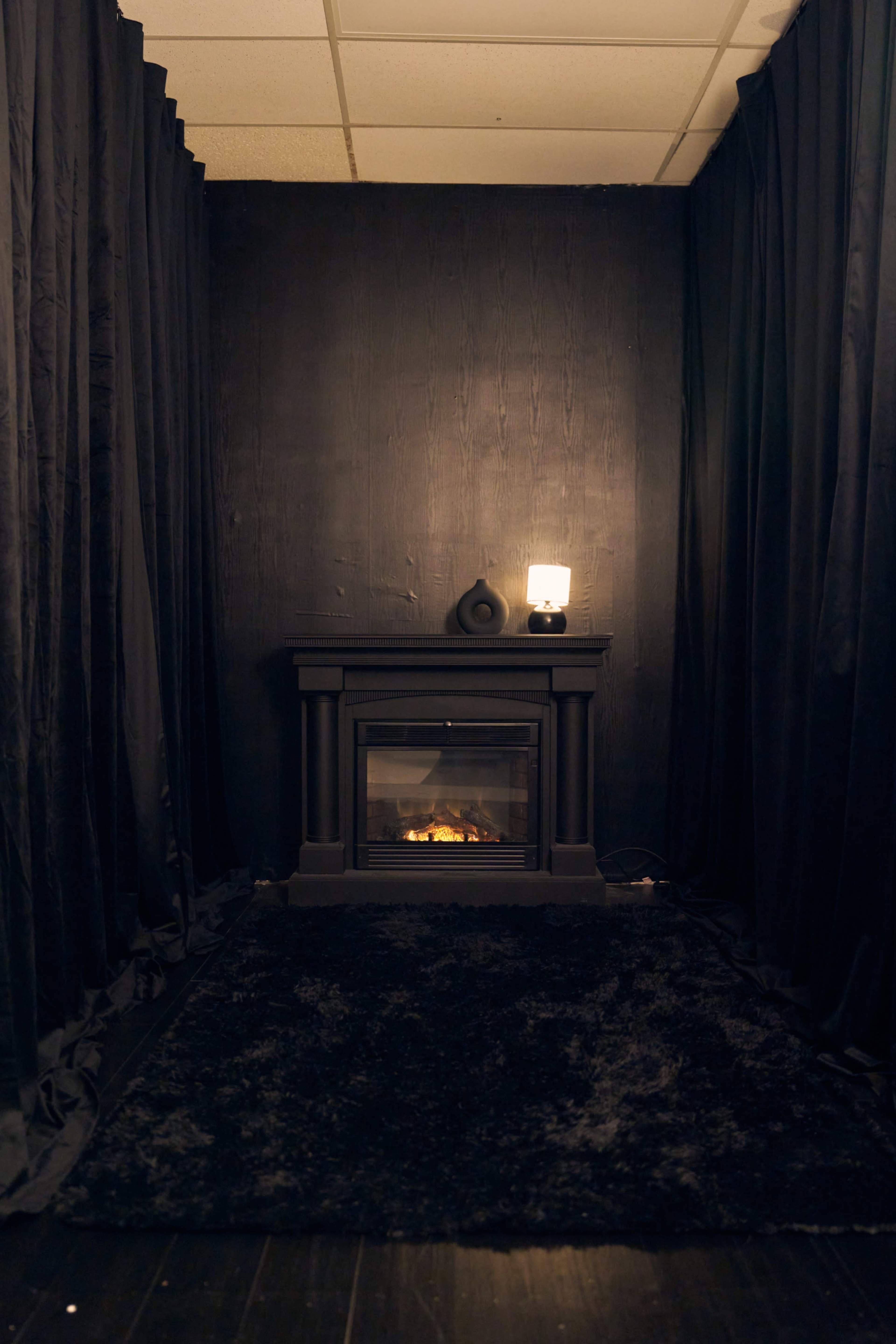 The image shows a dimly lit alcove featuring a black fireplace with an electric flame, surrounded by dark curtains and a rug.