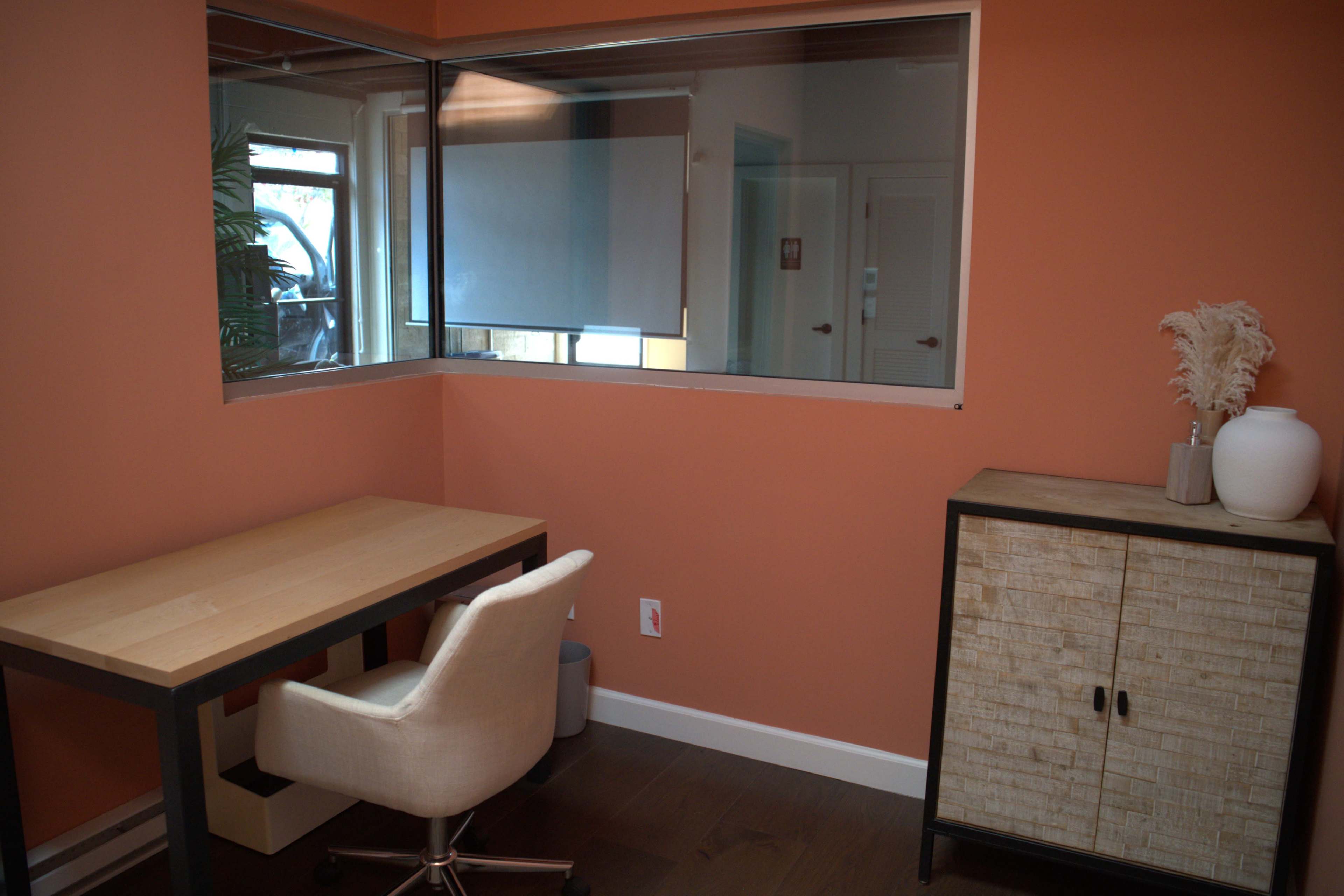 The image shows a small office space with a wooden desk and a white chair, against an orange-painted wall, featuring a glass window that overlooks another area.
