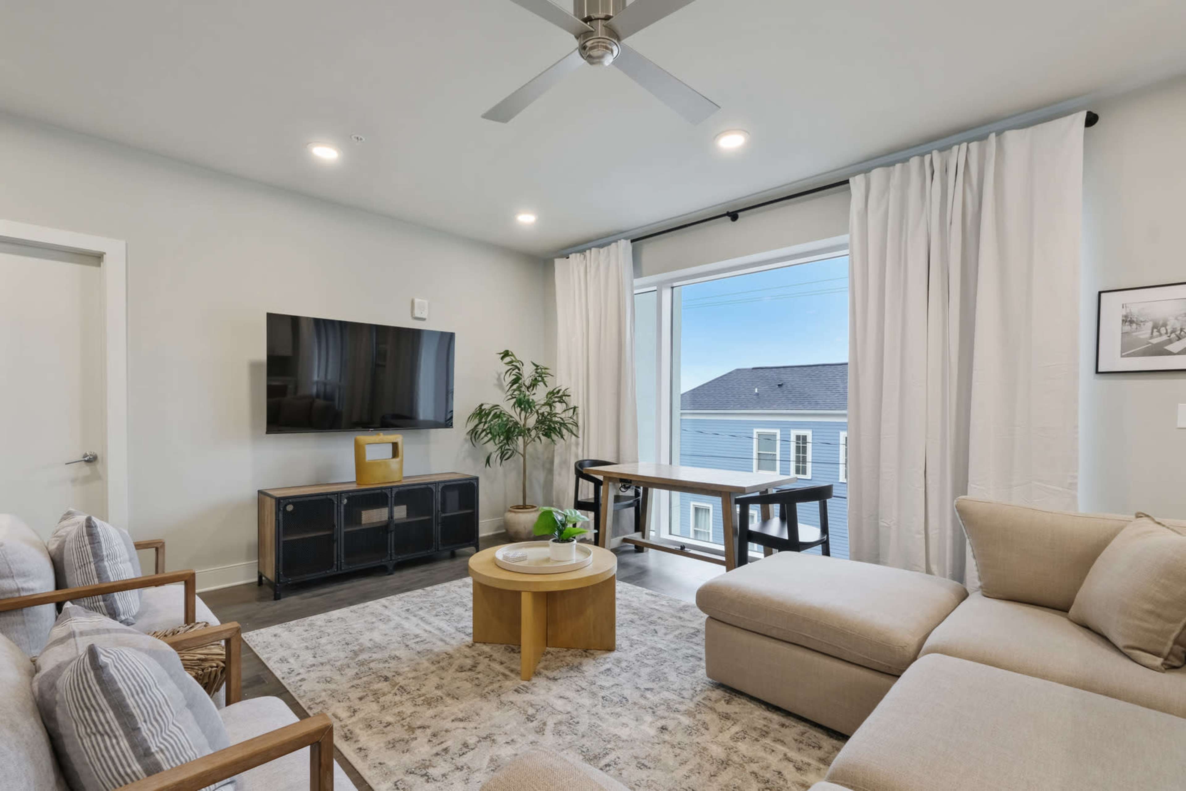 The image shows a modern living room with a sectional sofa, a wooden coffee table, a television mounted on the wall, and large windows with sheer curtains.