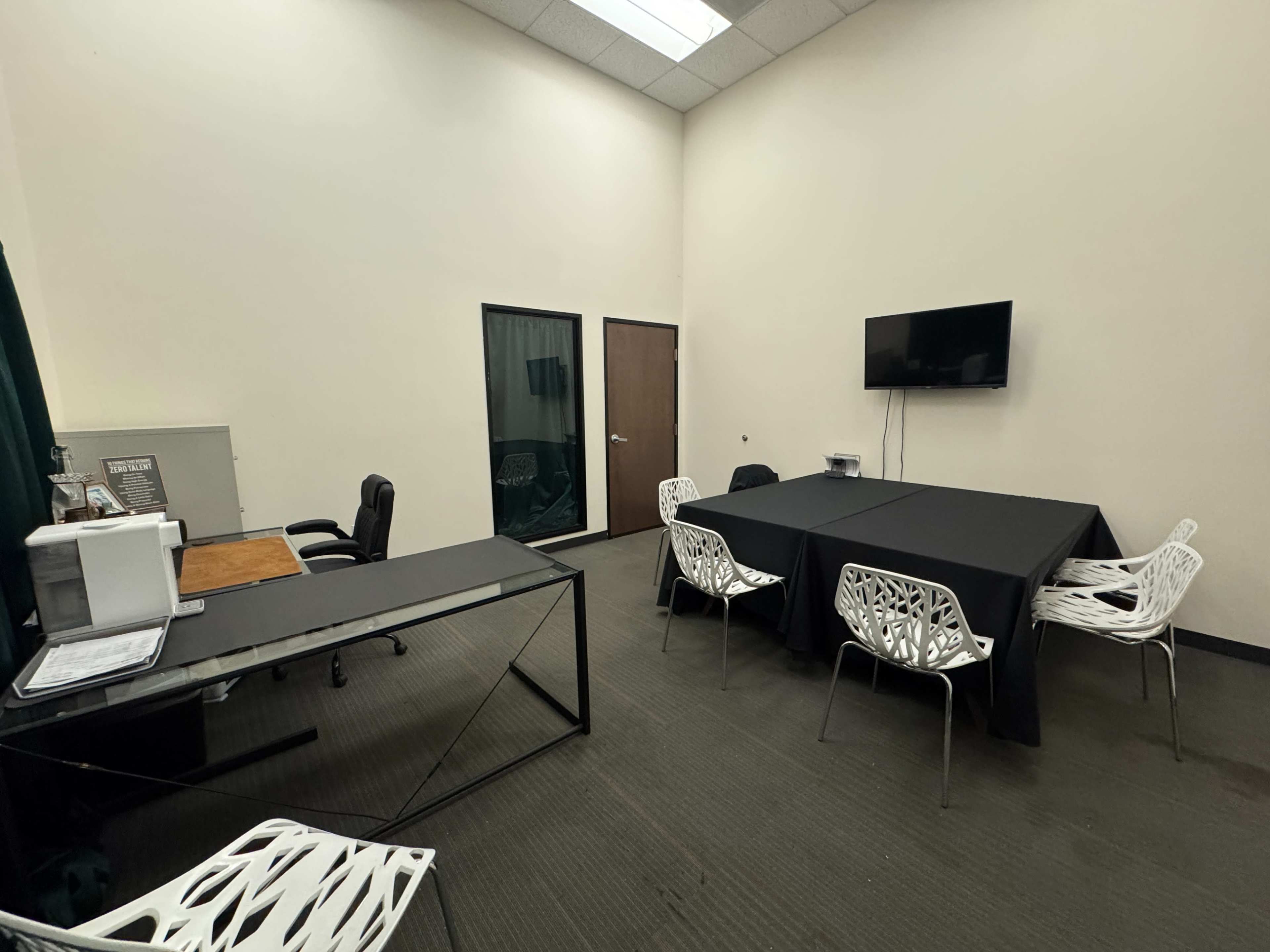 The image shows a spacious meeting room with a black table, several white chairs, a television mounted on the wall, and a desk with a chair in one corner.