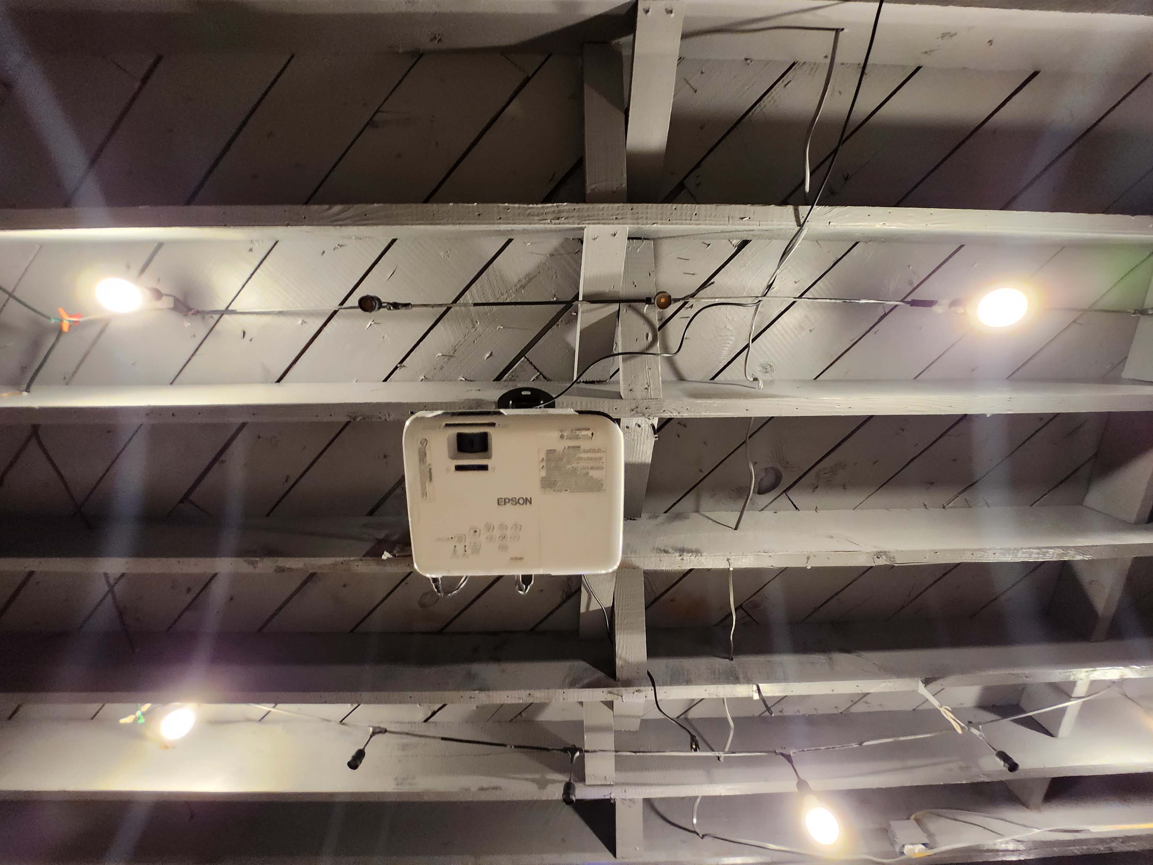 A ceiling-mounted Epson projector is installed on wooden beams with string lights overhead.