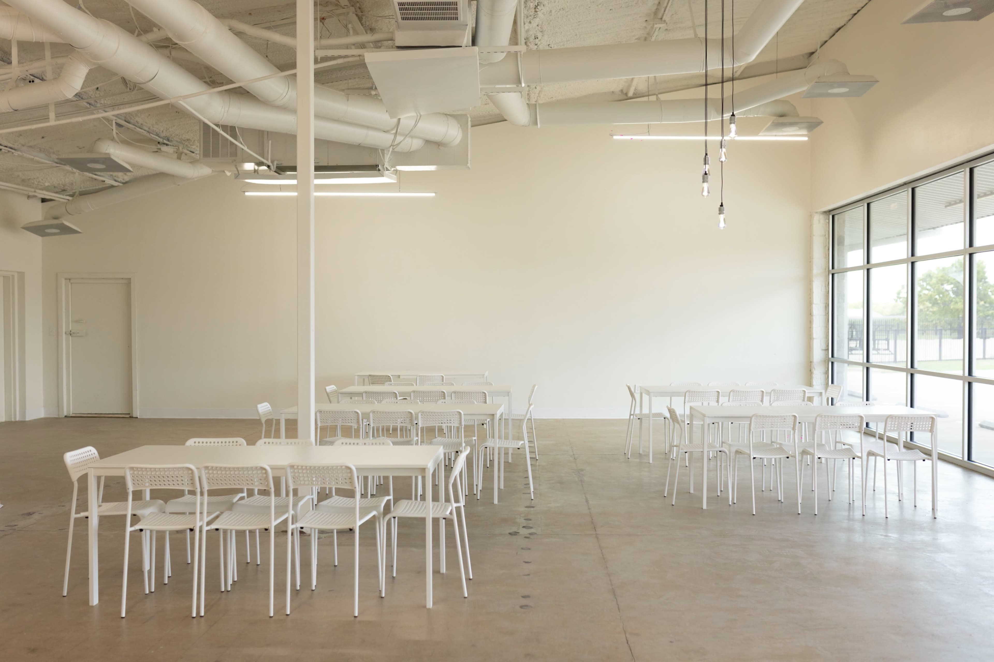 The image shows a spacious, minimalistic room with white walls and several white tables and chairs arranged neatly.