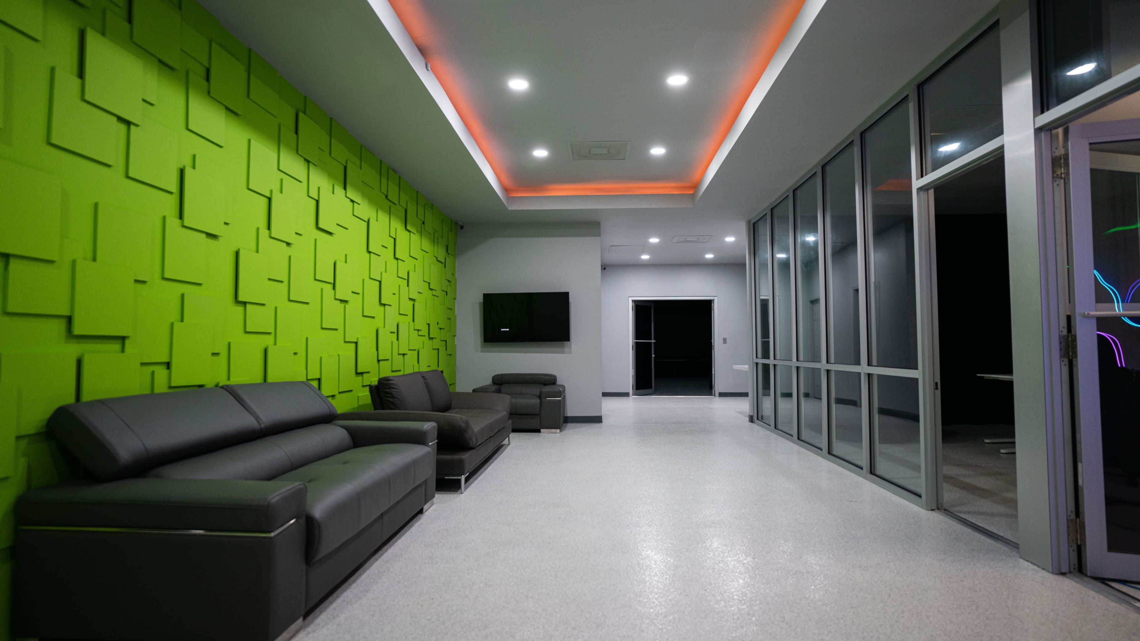 A modern hallway with a green accent wall, dark sofas, and a glass partition leading to another room.