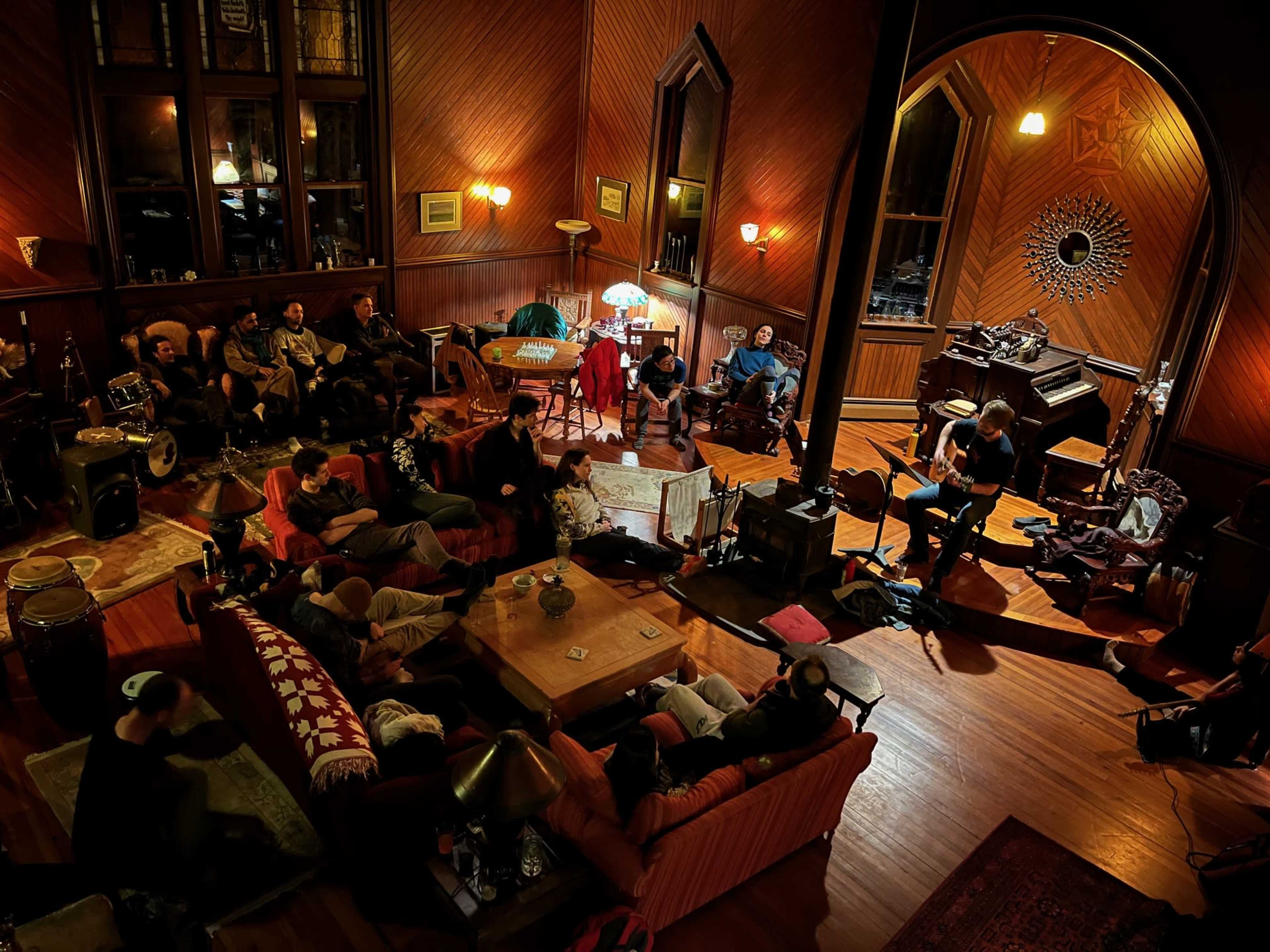A group of people gather in a warmly lit room with wooden walls, some seated on sofas and others engaged in conversation near a piano.