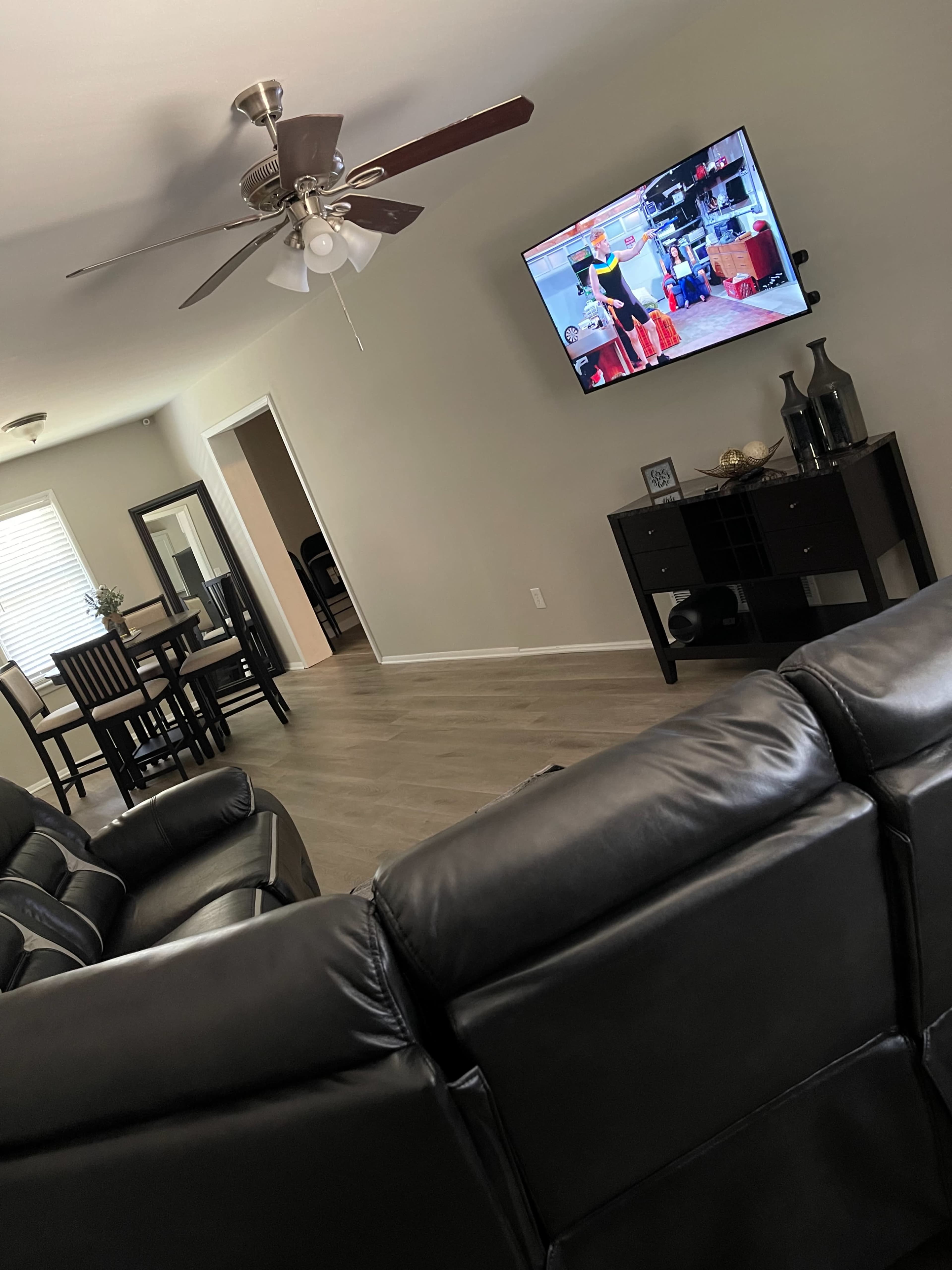 The living room features a black leather sectional sofa facing a television displaying a scene, with a dining area visible in the background.