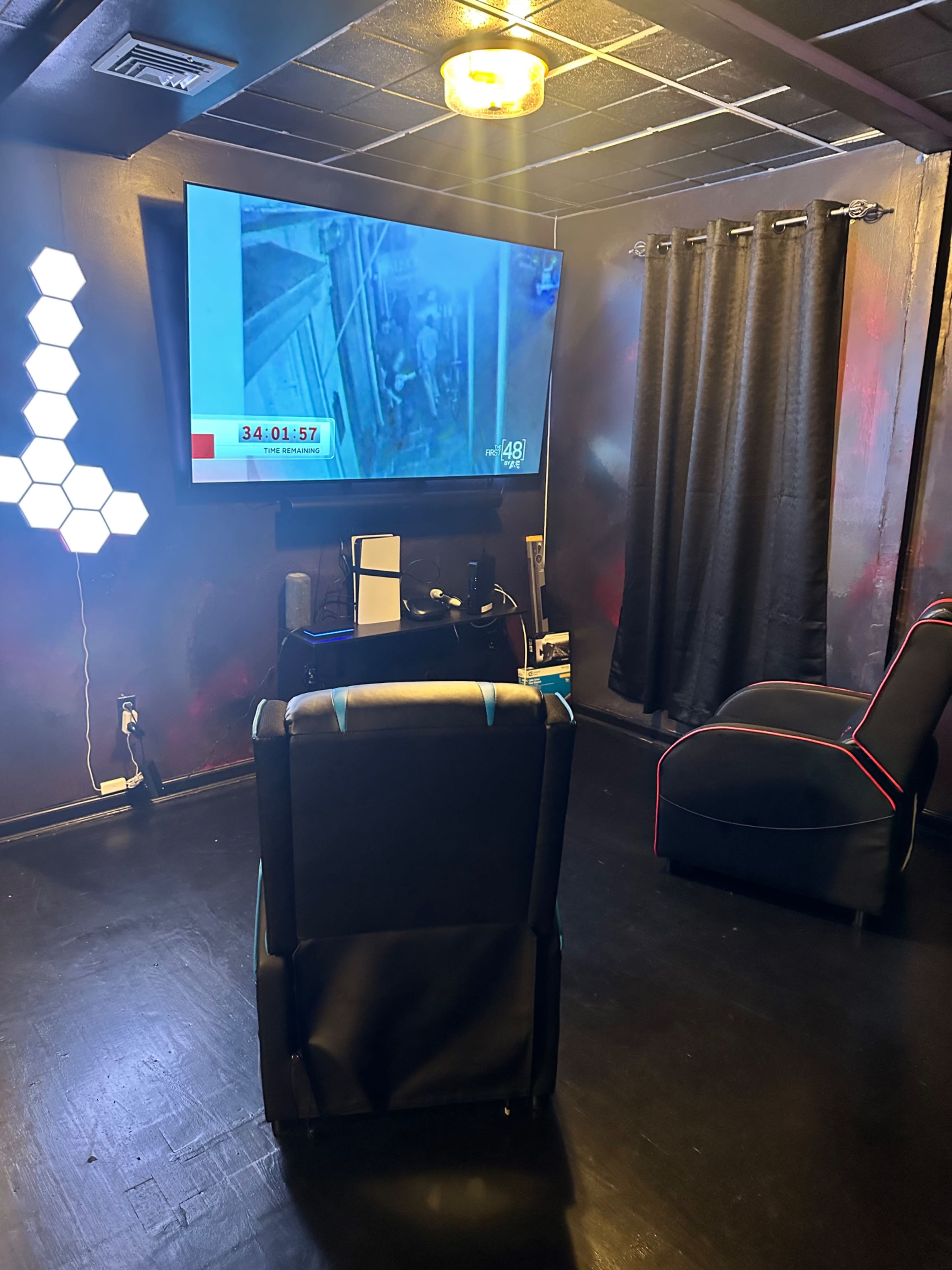 The image shows a dimly lit room with a large television displaying a timer, a gaming console on a small stand, and two chairs positioned for viewing.