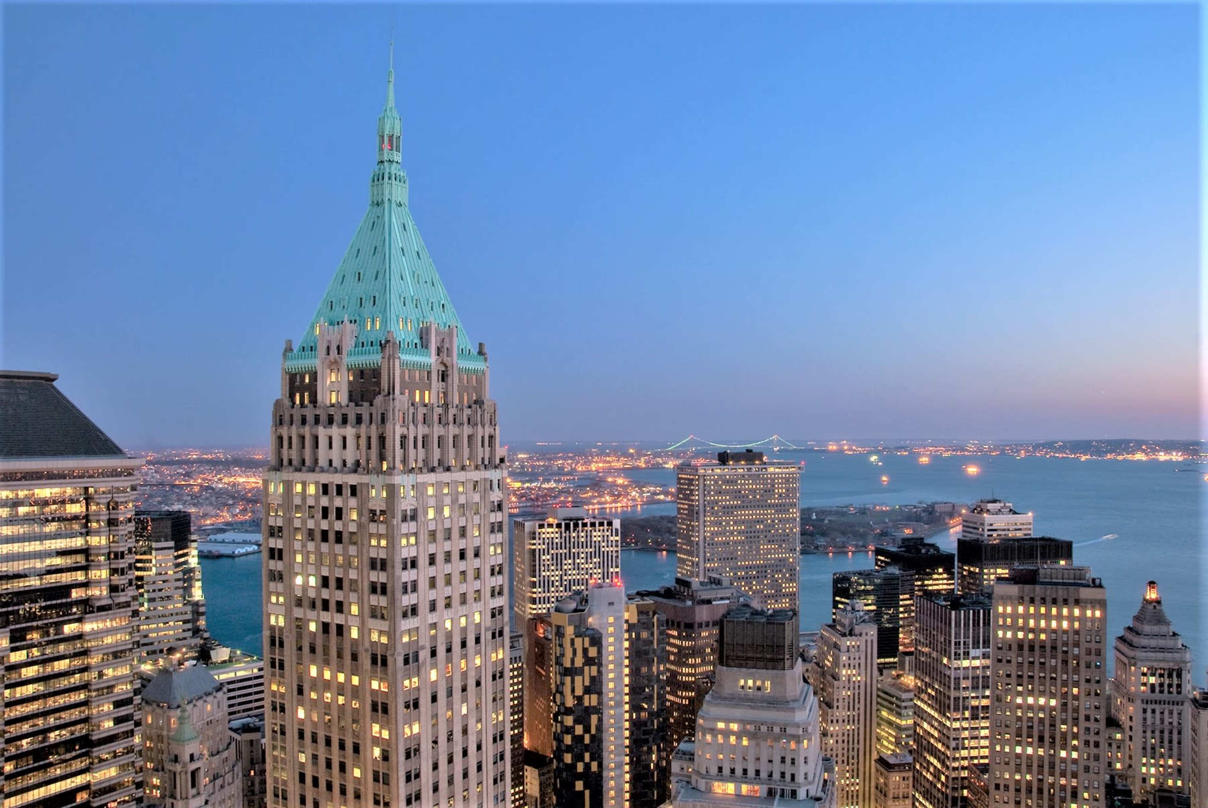 A panoramic view of a city skyline at dusk, highlighting buildings with a prominent green-tipped spire and distant bridges over water.