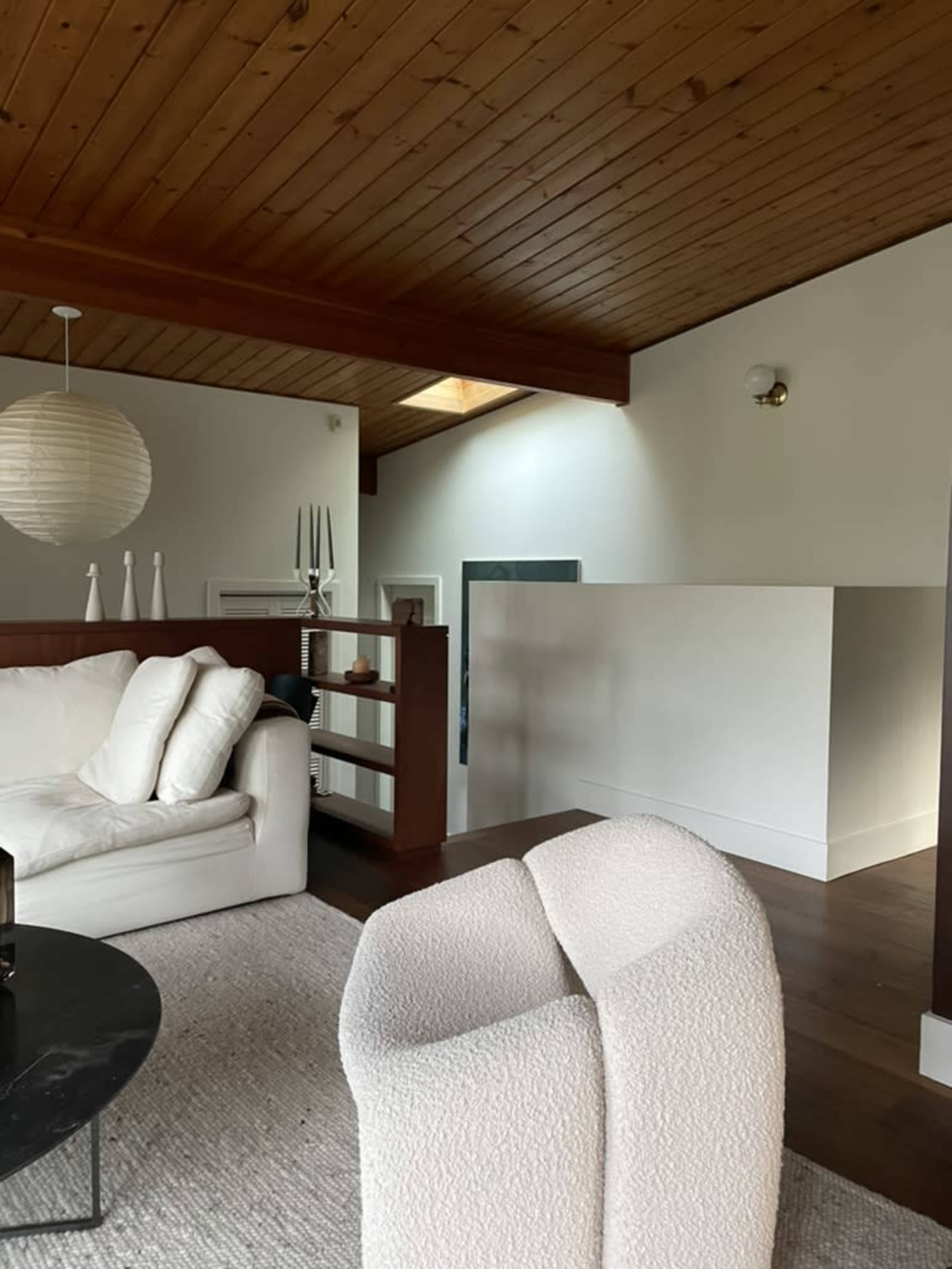 The image shows a living room with a white sofa, a circular table, and a textured chair, featuring wooden ceiling beams and a skylight.