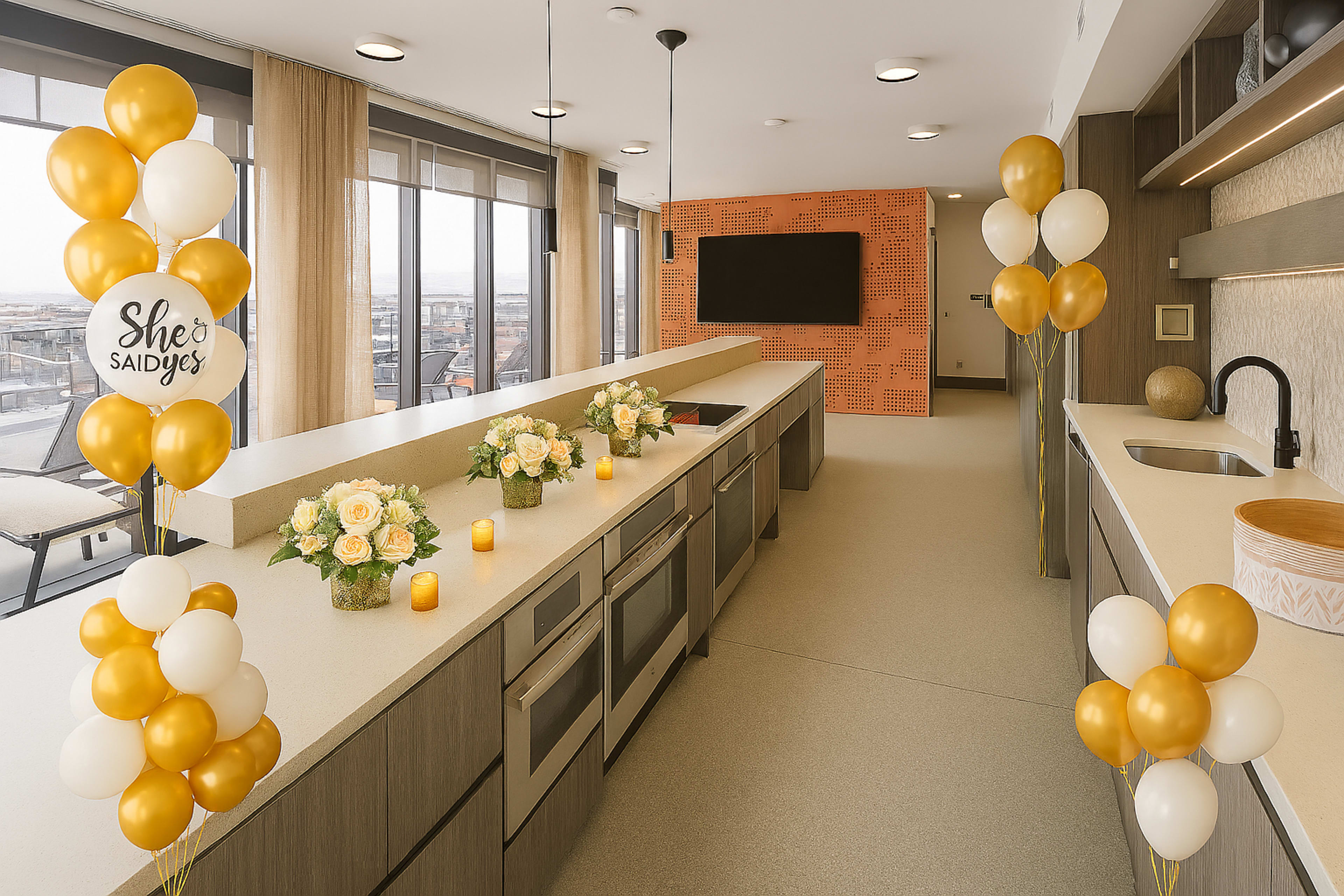 The image shows a modern kitchen with a long countertop decorated with yellow and white balloons, floral arrangements, and candles, set against a backdrop of large windows.