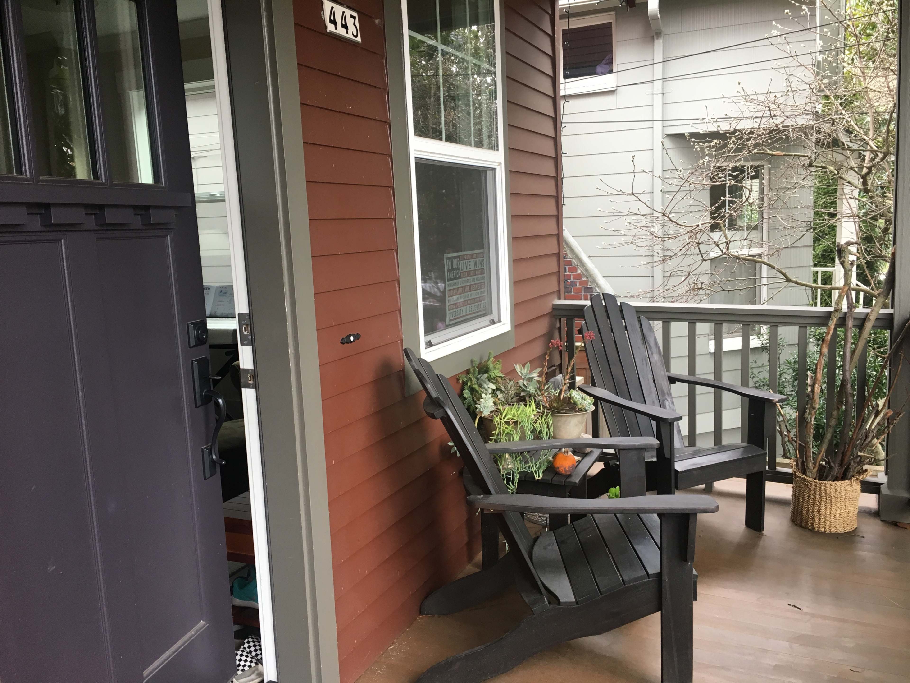A porch with two black wooden chairs, a small potted plant, and a house number displayed on the brown wall of the house.