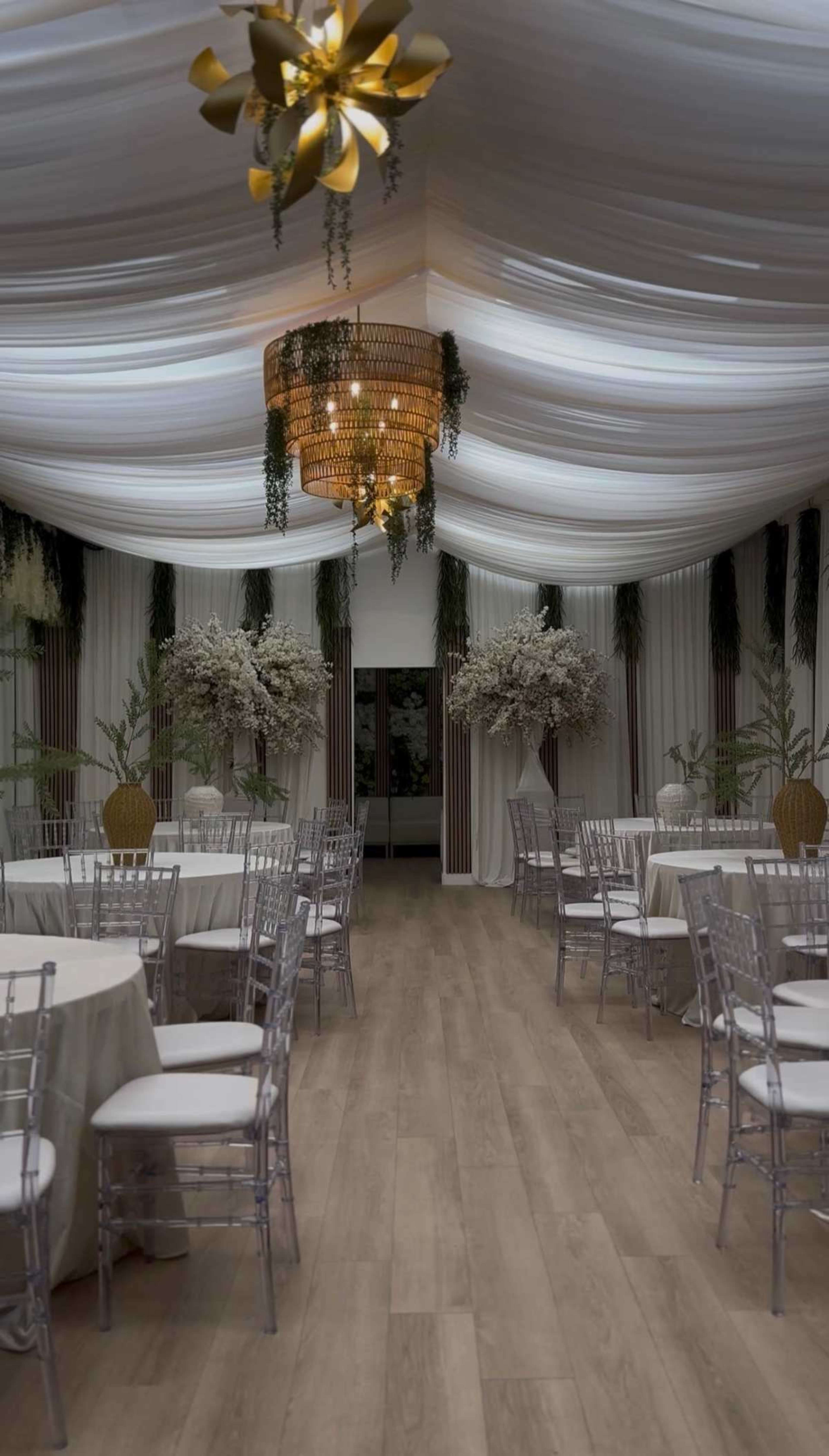 The image shows a beautifully decorated indoor venue with white draped ceilings, elegant chandeliers, and round tables arranged with clear chairs.