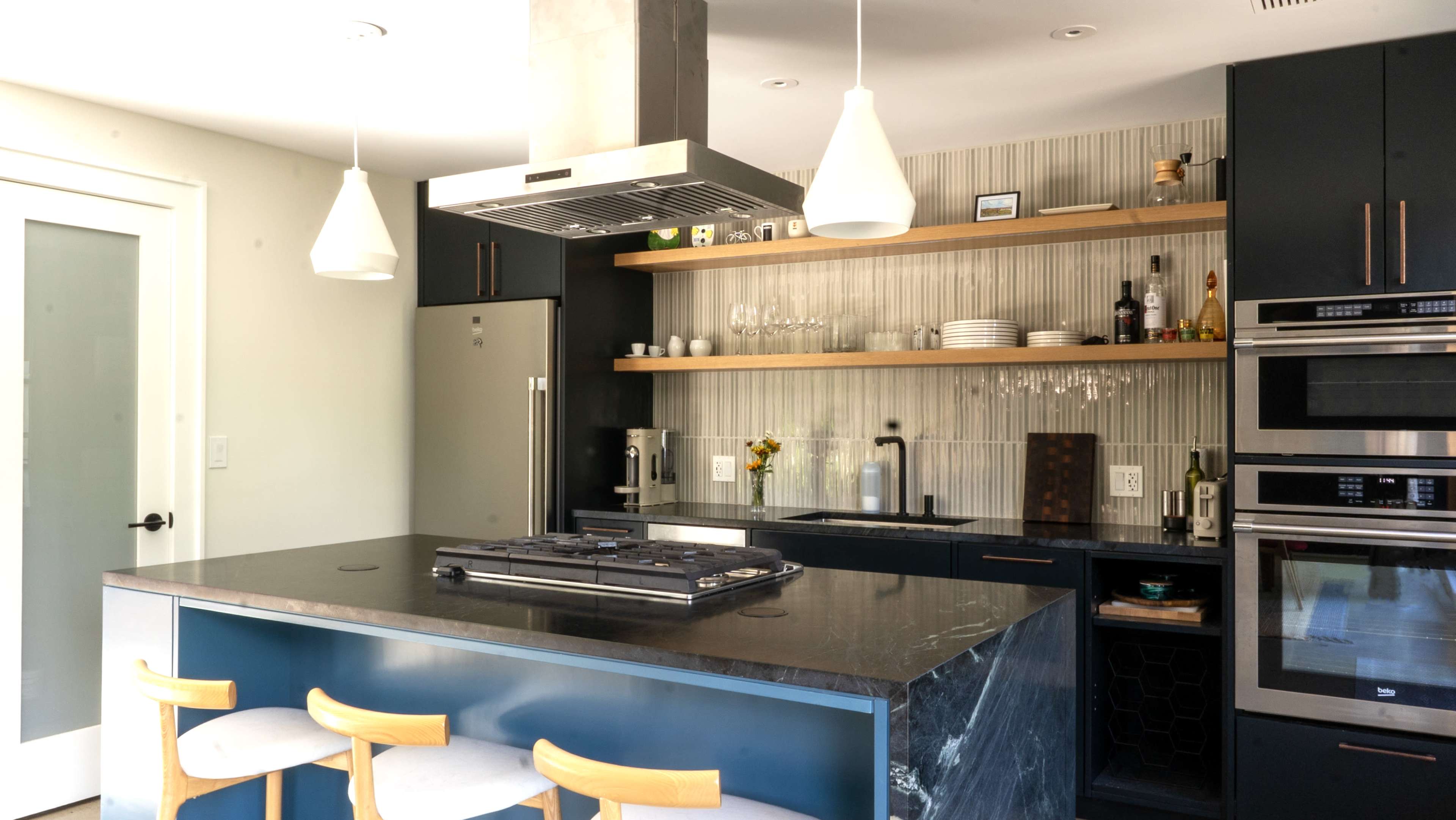 The image shows a modern kitchen featuring dark cabinetry, a large island with a cooktop, and open shelving displaying various kitchen items.