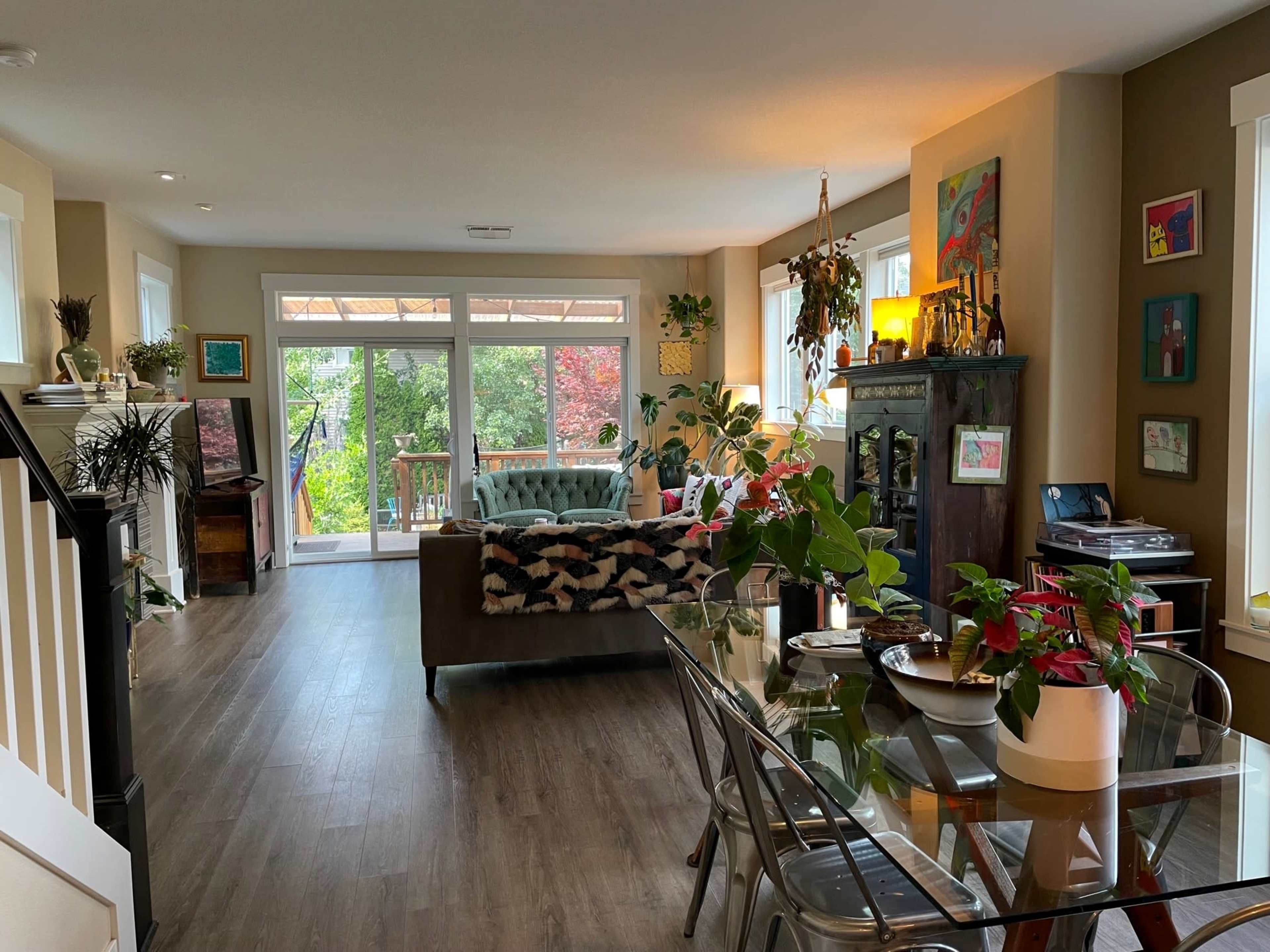 A well-lit living room features a green sofa, a dining table with plants, and large windows overlooking a deck and garden.