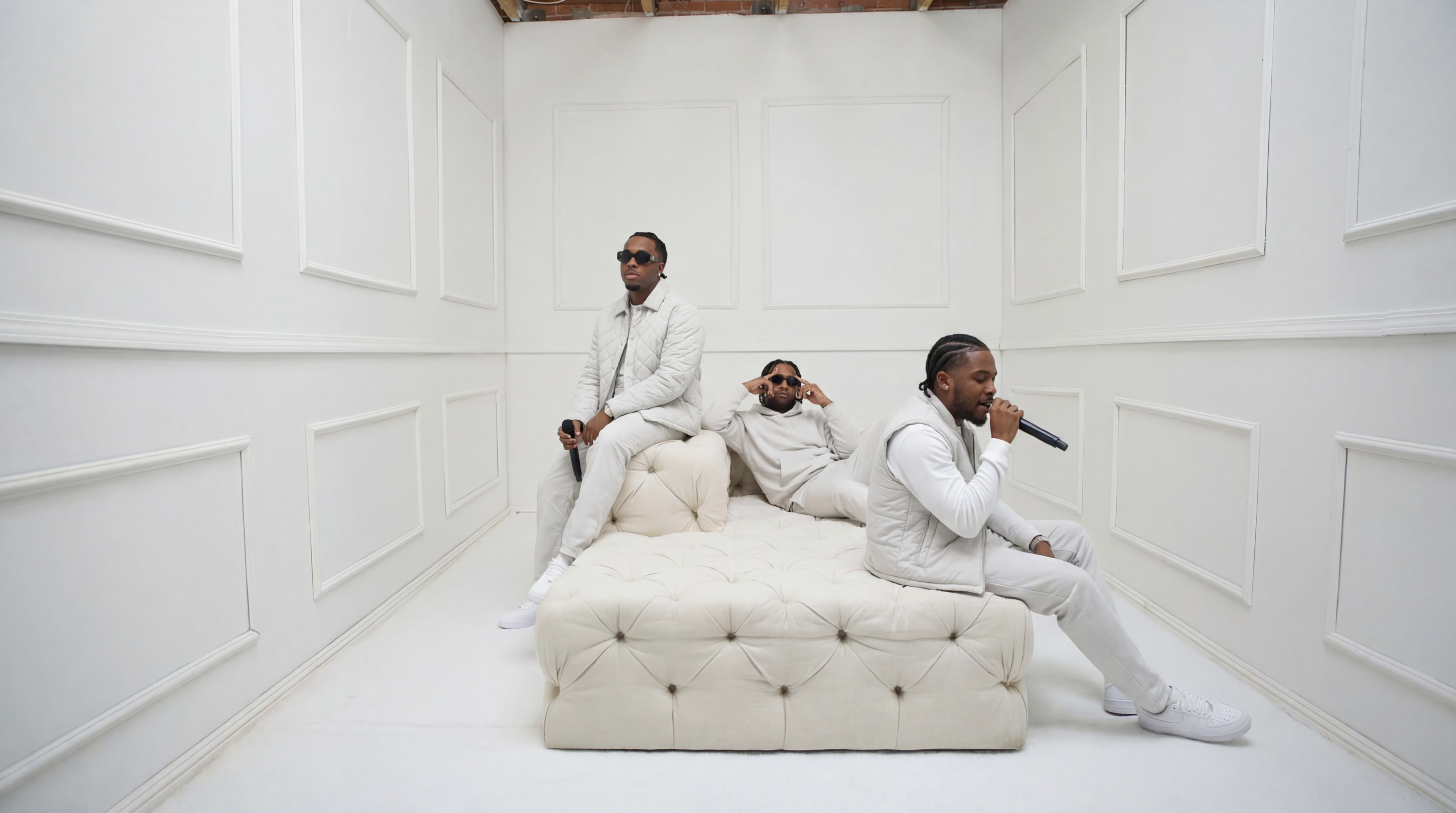 Three men dressed in white are performing and posing on a white tufted sofa in a minimalist, all-white room.