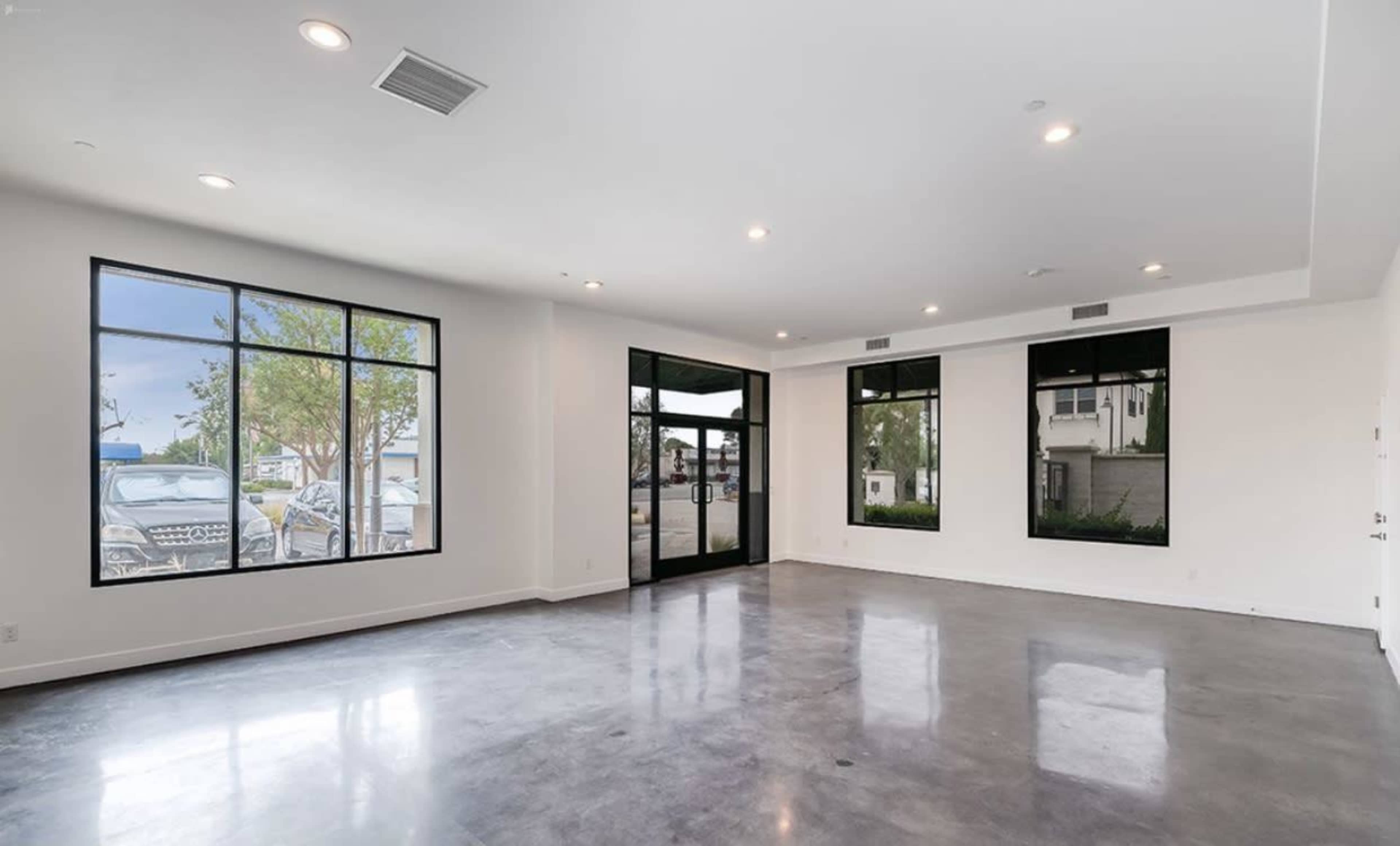 The image shows an empty commercial space with large windows casting natural light onto a polished concrete floor.