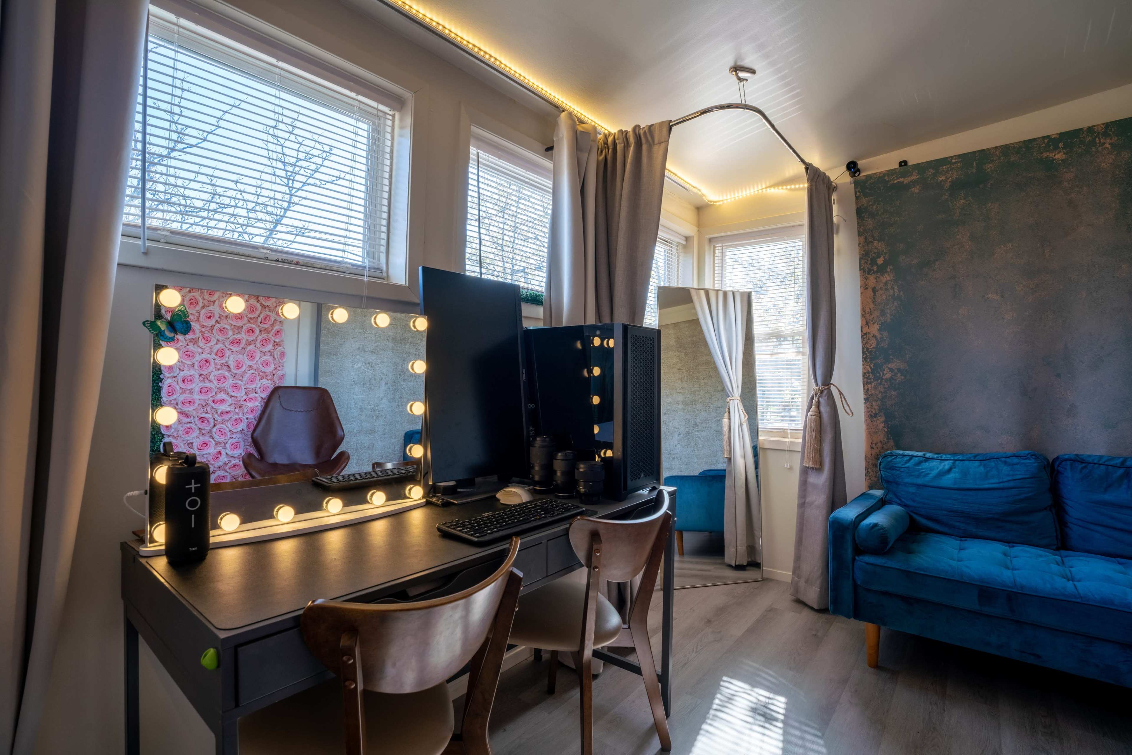 A bright room with a desk featuring a large mirror surrounded by lights, a computer monitor, and a blue sofa, complemented by partially drawn curtains.