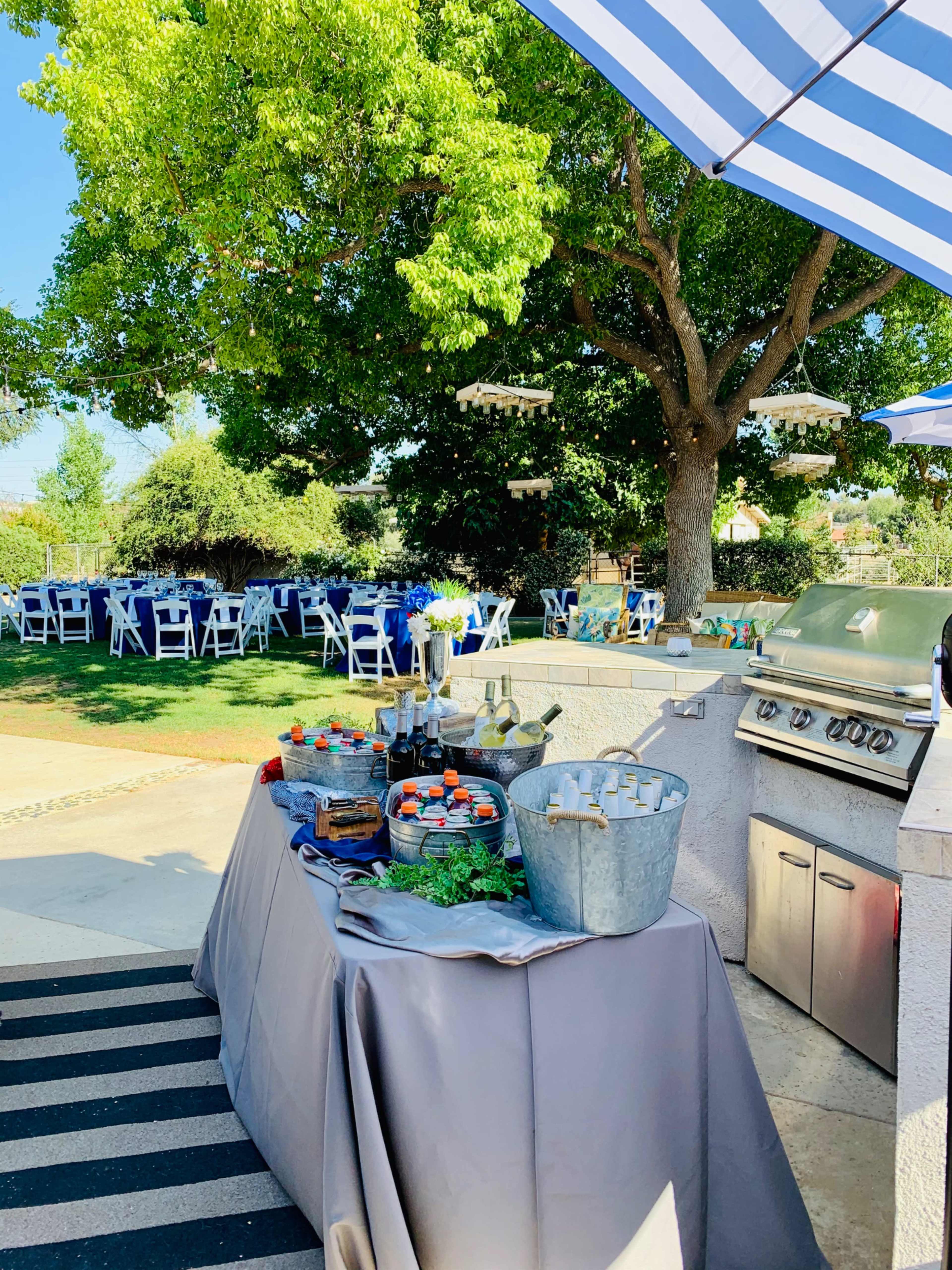 A buffet table is set up outdoors next to a grill, with seating arranged on a lawn shaded by a large tree and decorated with hanging lanterns.