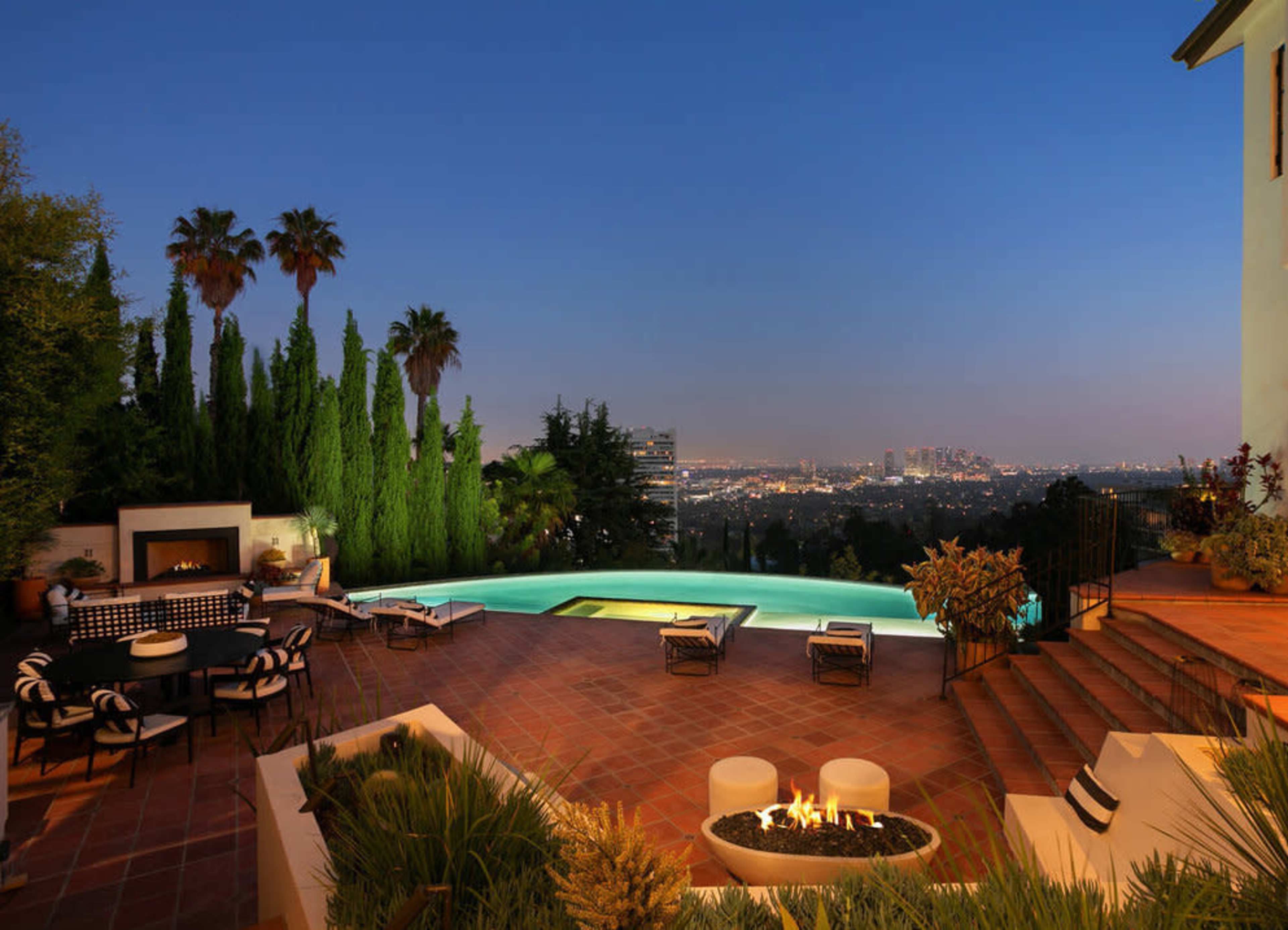 A luxury patio with a pool overlooks a city skyline, surrounded by palm trees and landscaped greenery, illuminated by evening light.