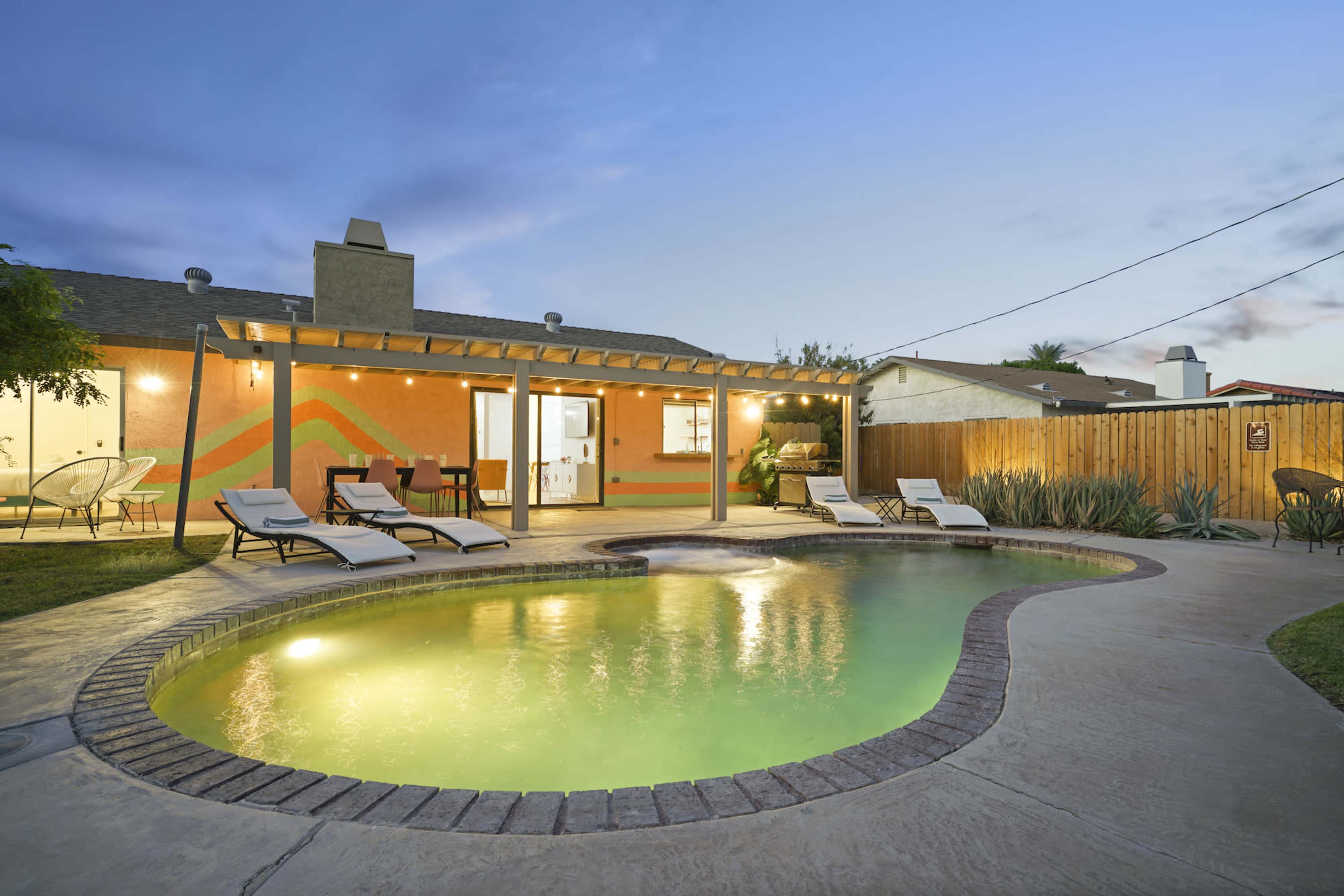 A backyard featuring a swimming pool with lounge chairs, a covered patio area, and vibrant wall art at dusk.