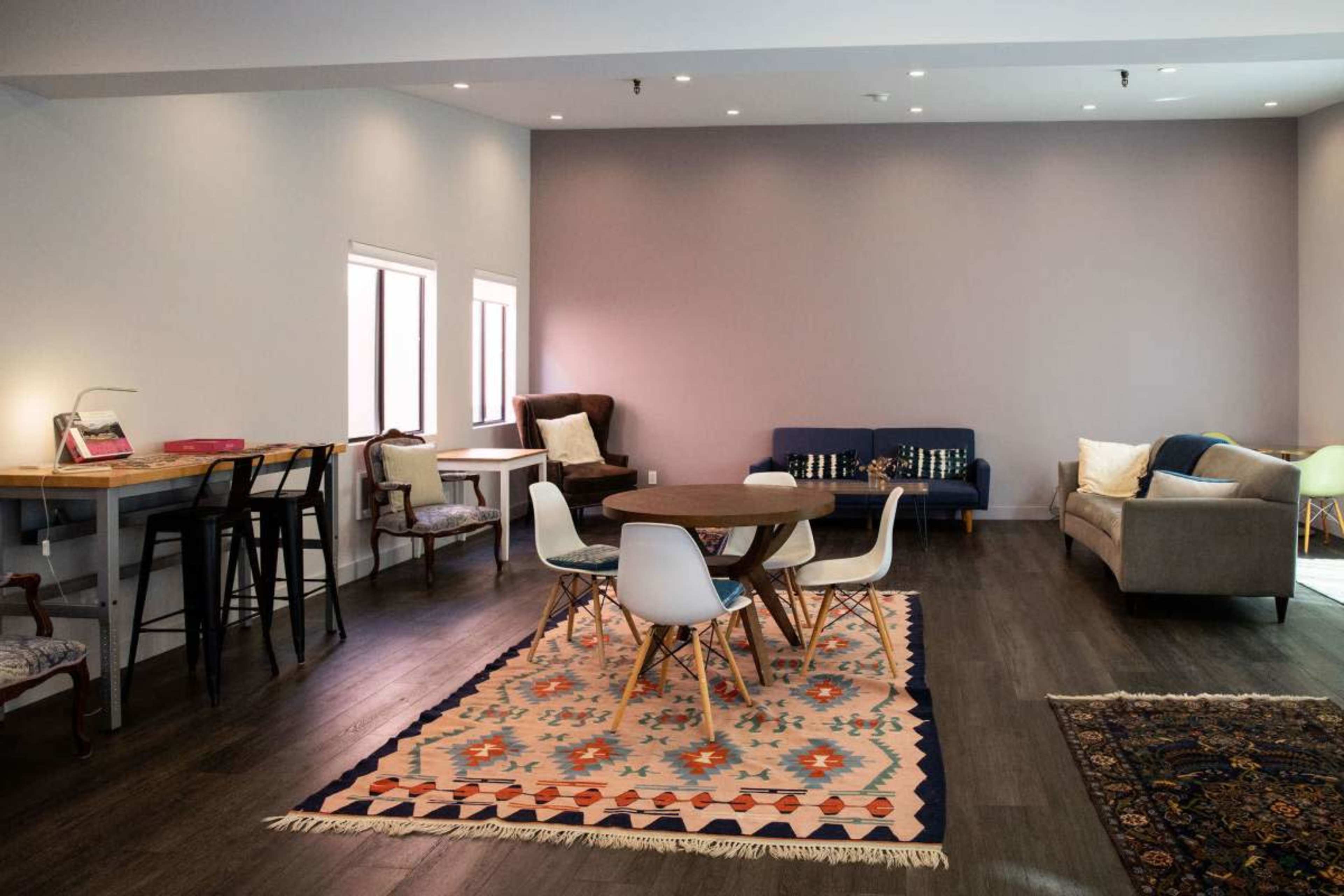 The image shows a spacious, modern room with various seating arrangements, including a round dining table surrounded by chairs, two sofas, and several small tables on a wooden floor.