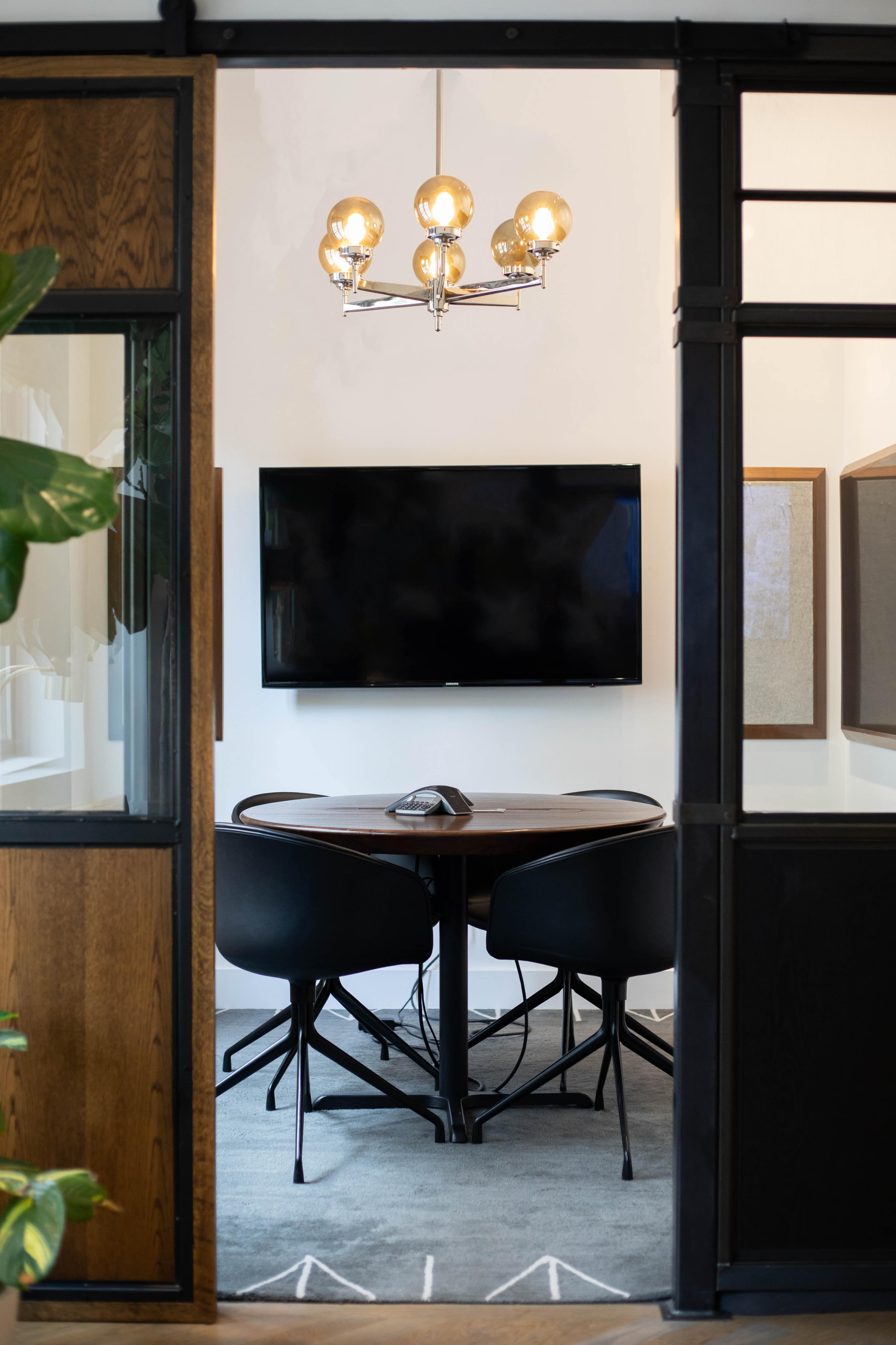 A modern meeting room features a round table with two chairs, a large wall-mounted TV, and a stylish light fixture above.