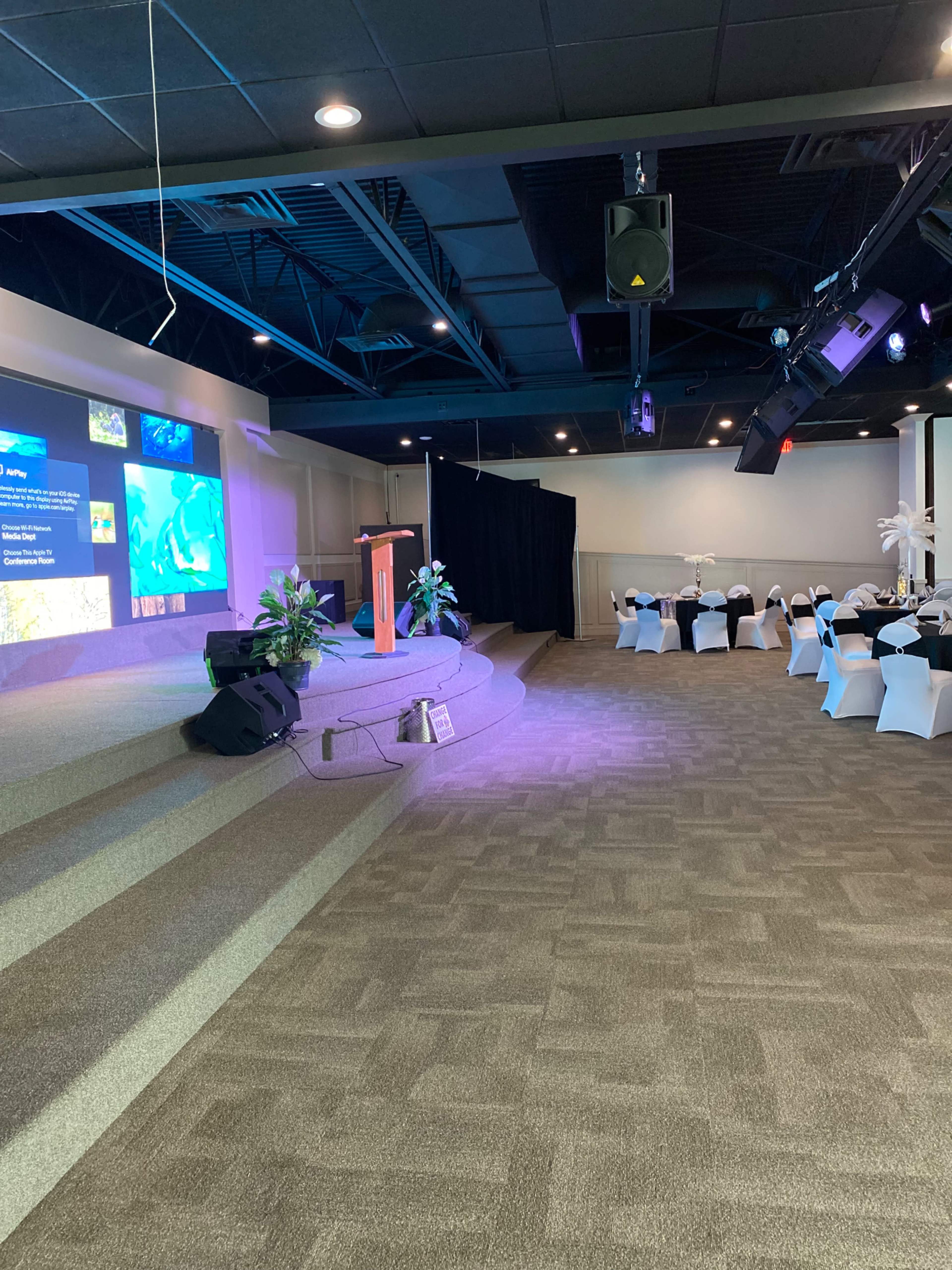 A stage with a backdrop featuring digital displays is set up in a banquet hall, with chairs arranged at tables nearby.