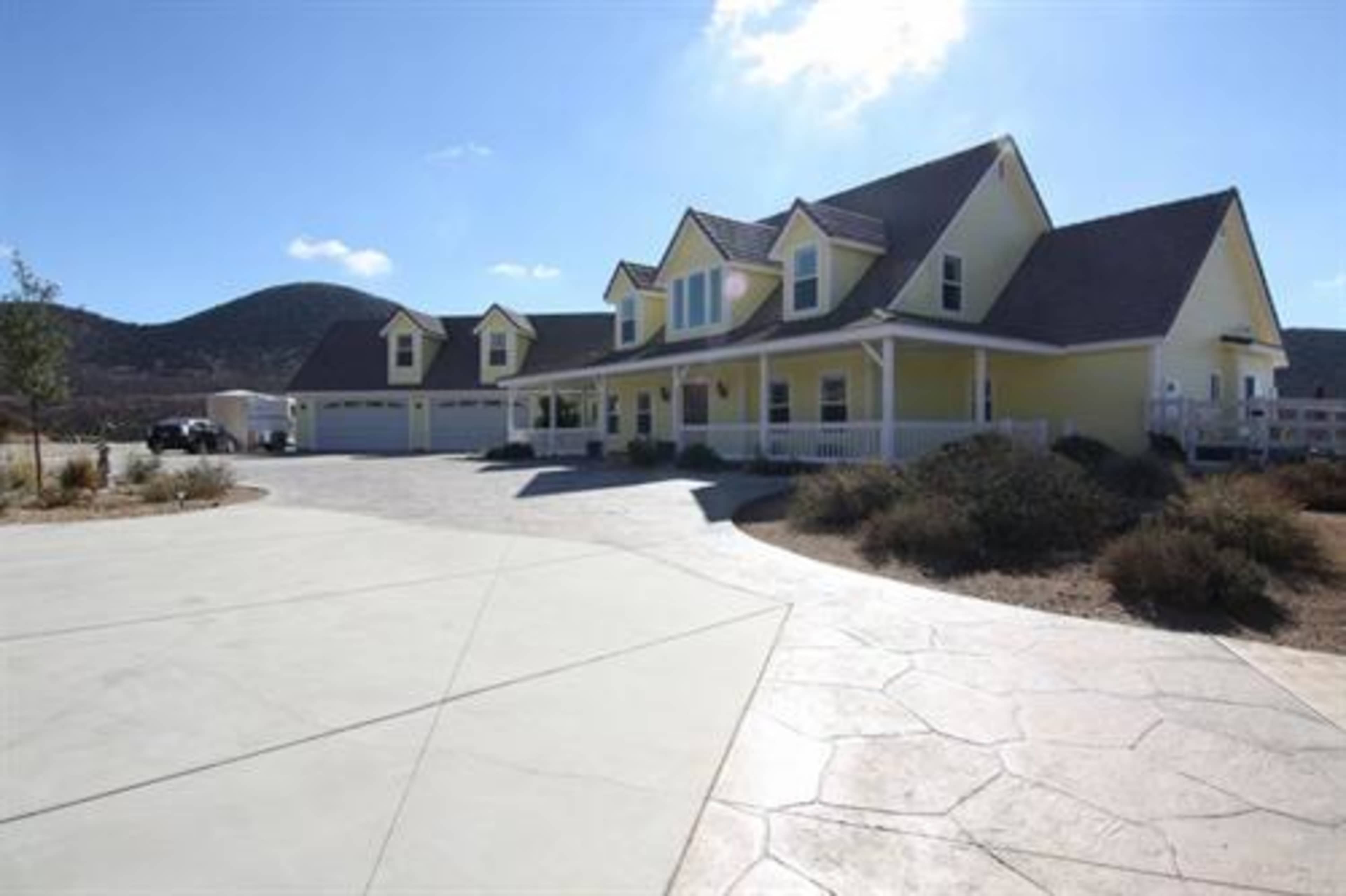 A large yellow house with multiple gabled roofs and a wide driveway is set against a mountainous background.