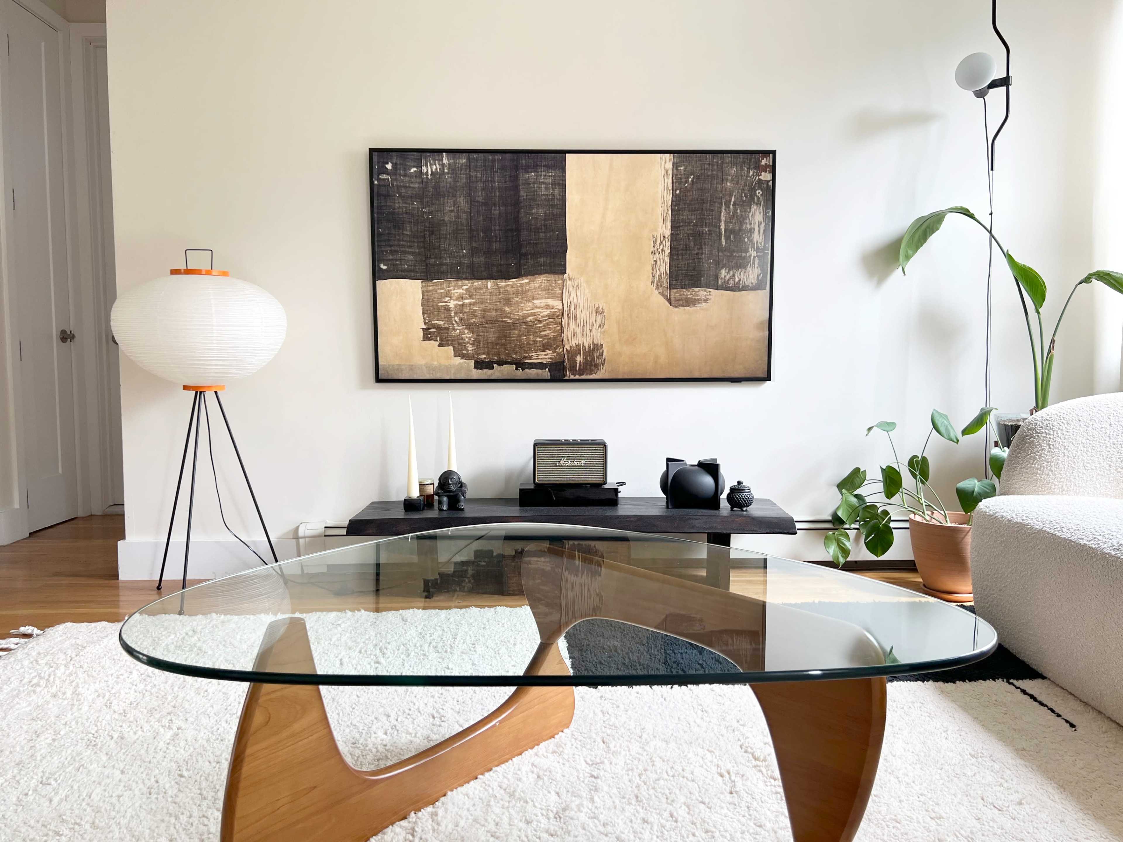 A modern living room with a glass coffee table, a wooden media console, a framed artwork on the wall, and plants in natural light.